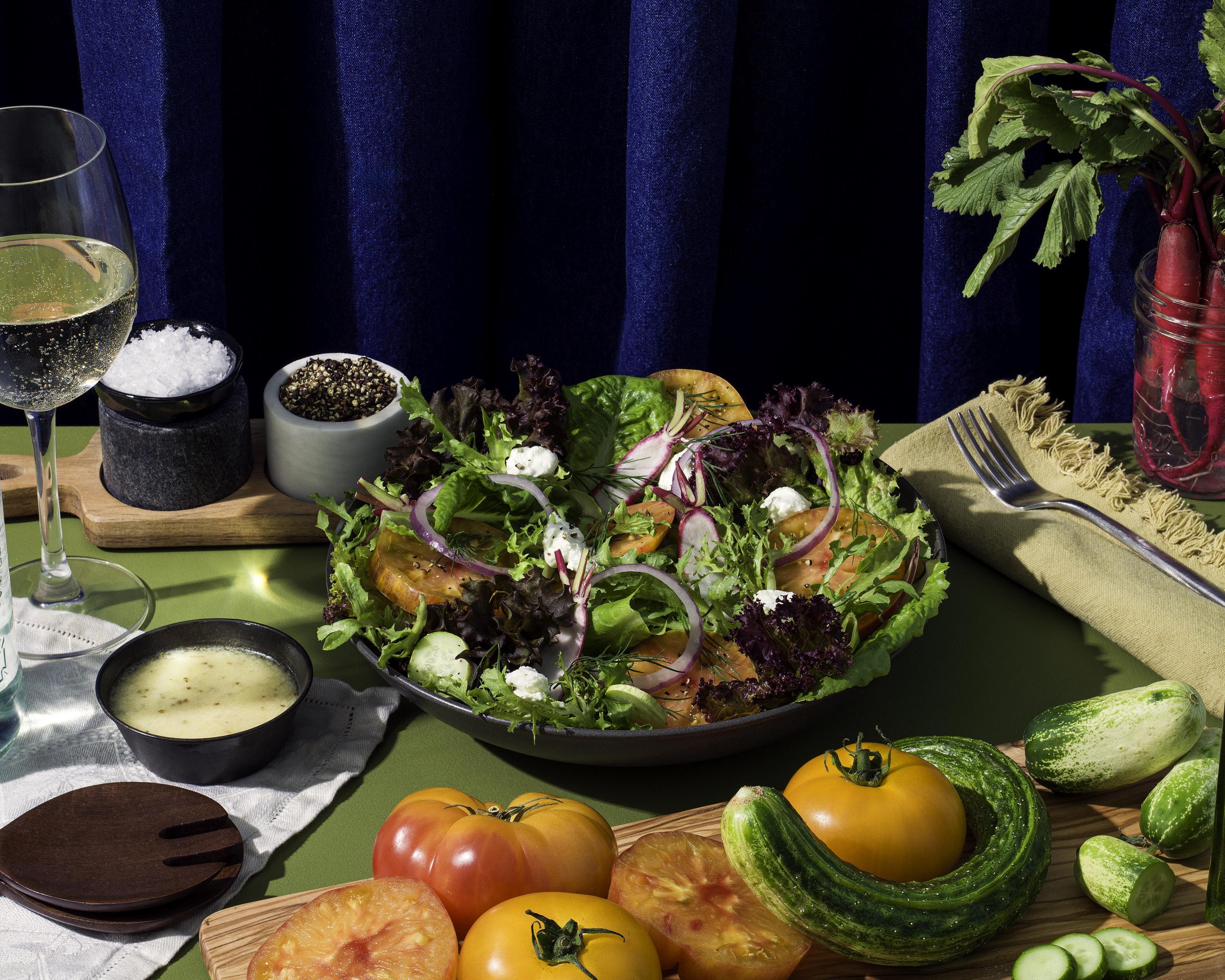 A colorful salad with tomatoes, cucumbers, onions, greens, and crumbled cheese on a table with vegetables, a glass of white wine, and condiments.