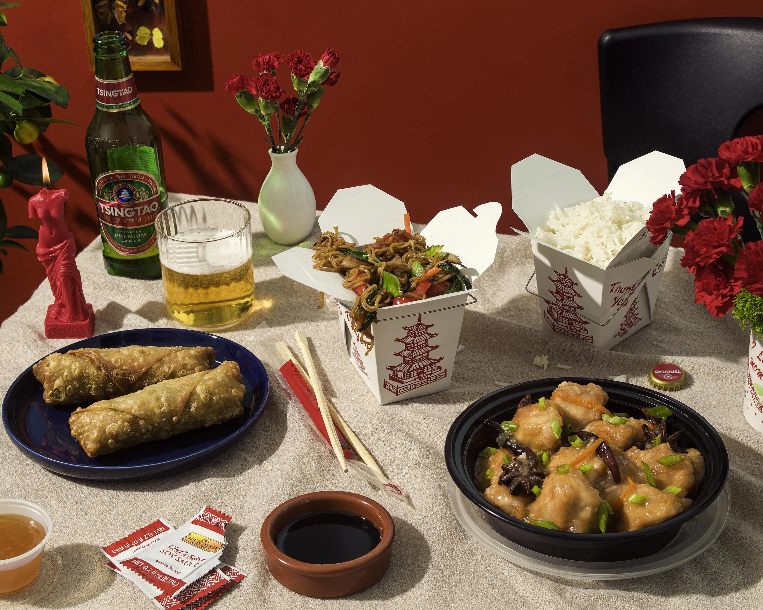 A table set with Chinese takeout food including egg rolls,  orange chicken, vegetable lo mein, Tsingtao beer, soy sauce,, chopsticks, a pink candle, and decorative flowers against a red background.