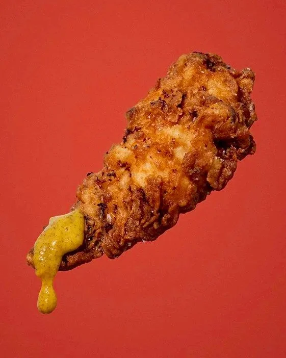 A fried chicken tender with a small amount of honey mustard on the tip, set against a plain red background.