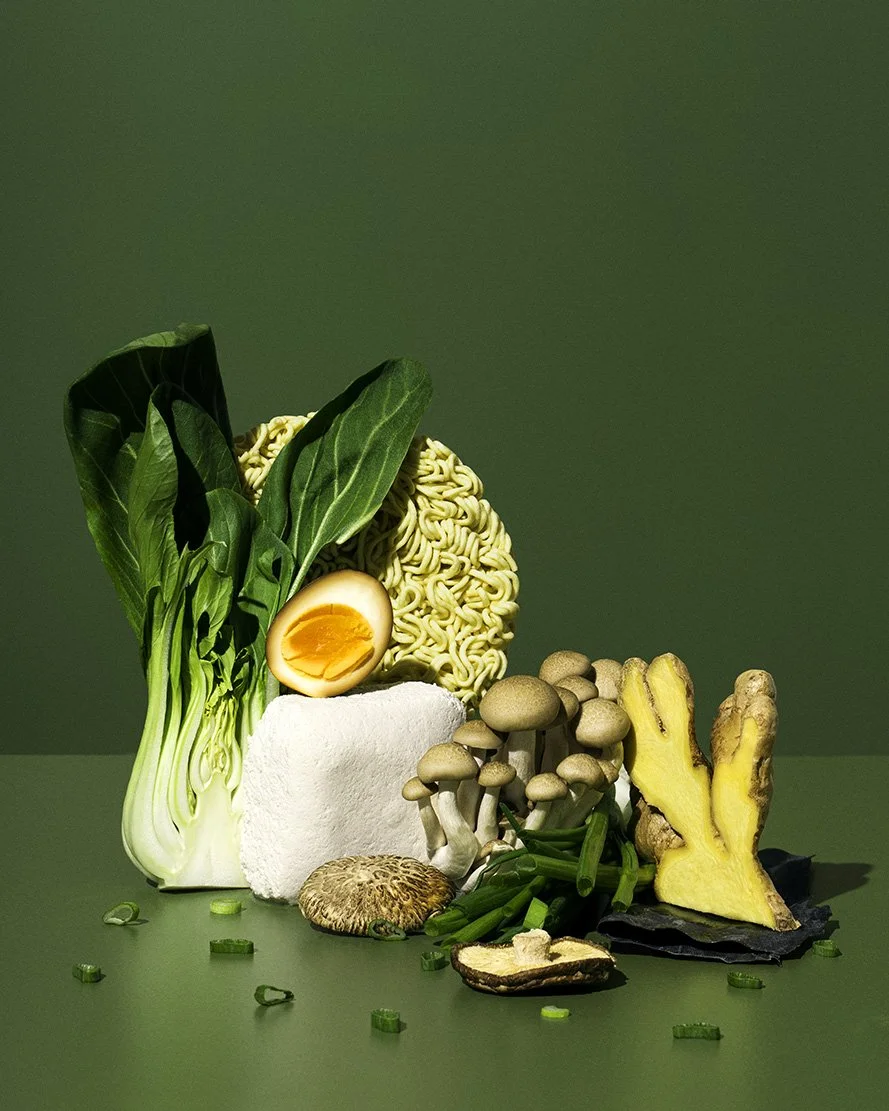 Arrangement of various fresh vegetables and instant ramen noodles on a green background.