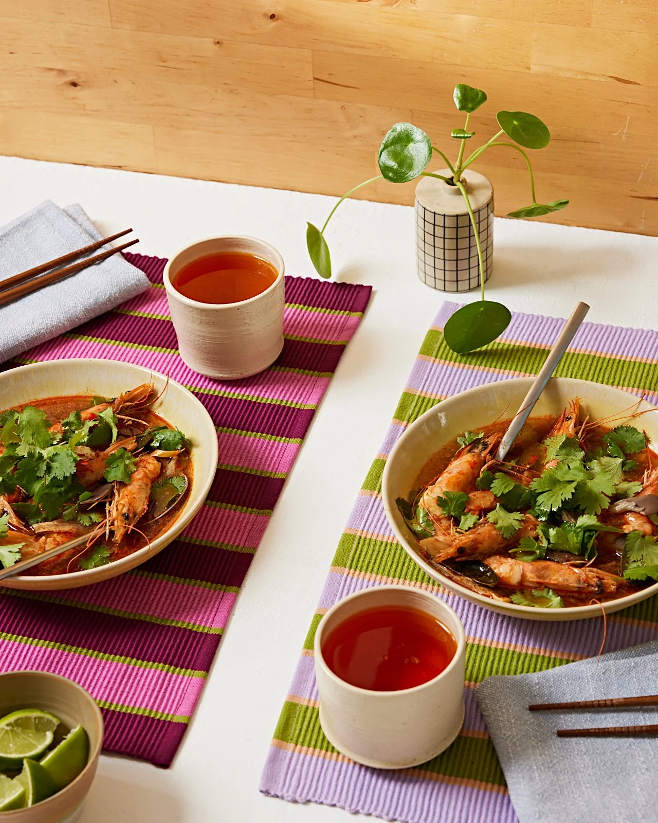 Two bowls of shrimp Tom Yum and herbs on colorful striped placemats, with cups of tea, a small bowl of lime wedges, a potted plant, and chopsticks on a white table.