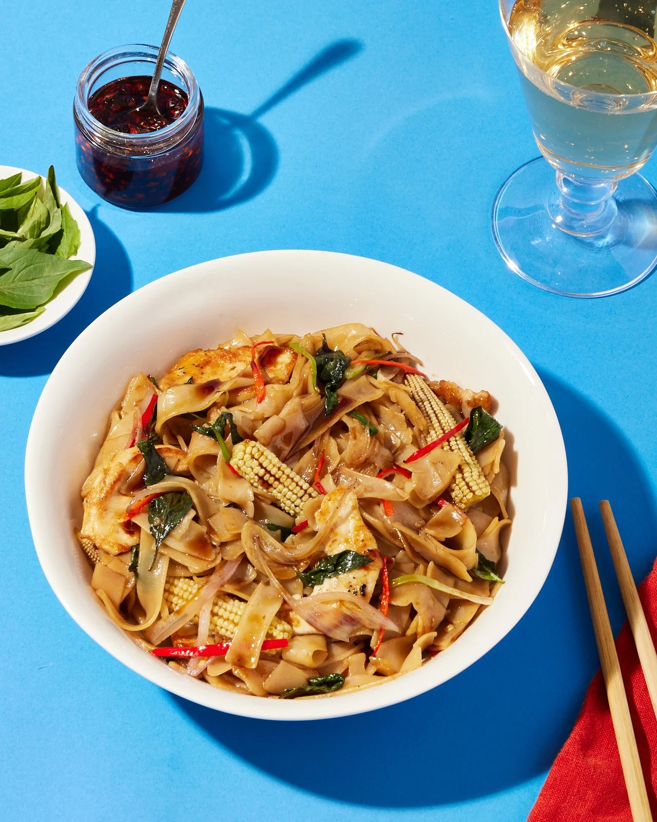 A bowl of stir-fried noodles with vegetables on a blue table, with a glass of white wine, a small jar of red sauce, and a small bowl of leafy greens nearby.