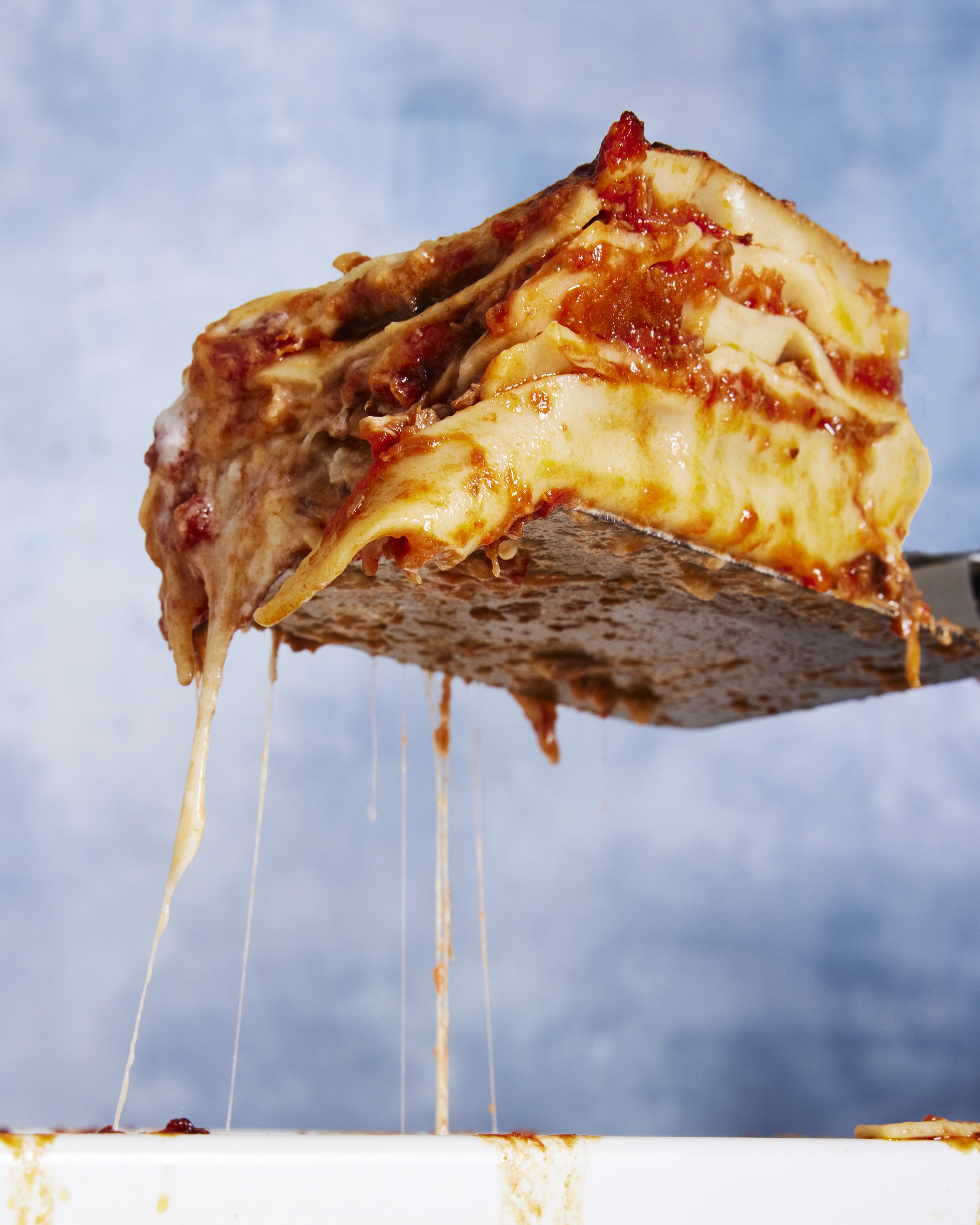 A slice of Lasagna with cheese stretching as it is lifted from a casserole dish, with a cloudy blue sky in the background.