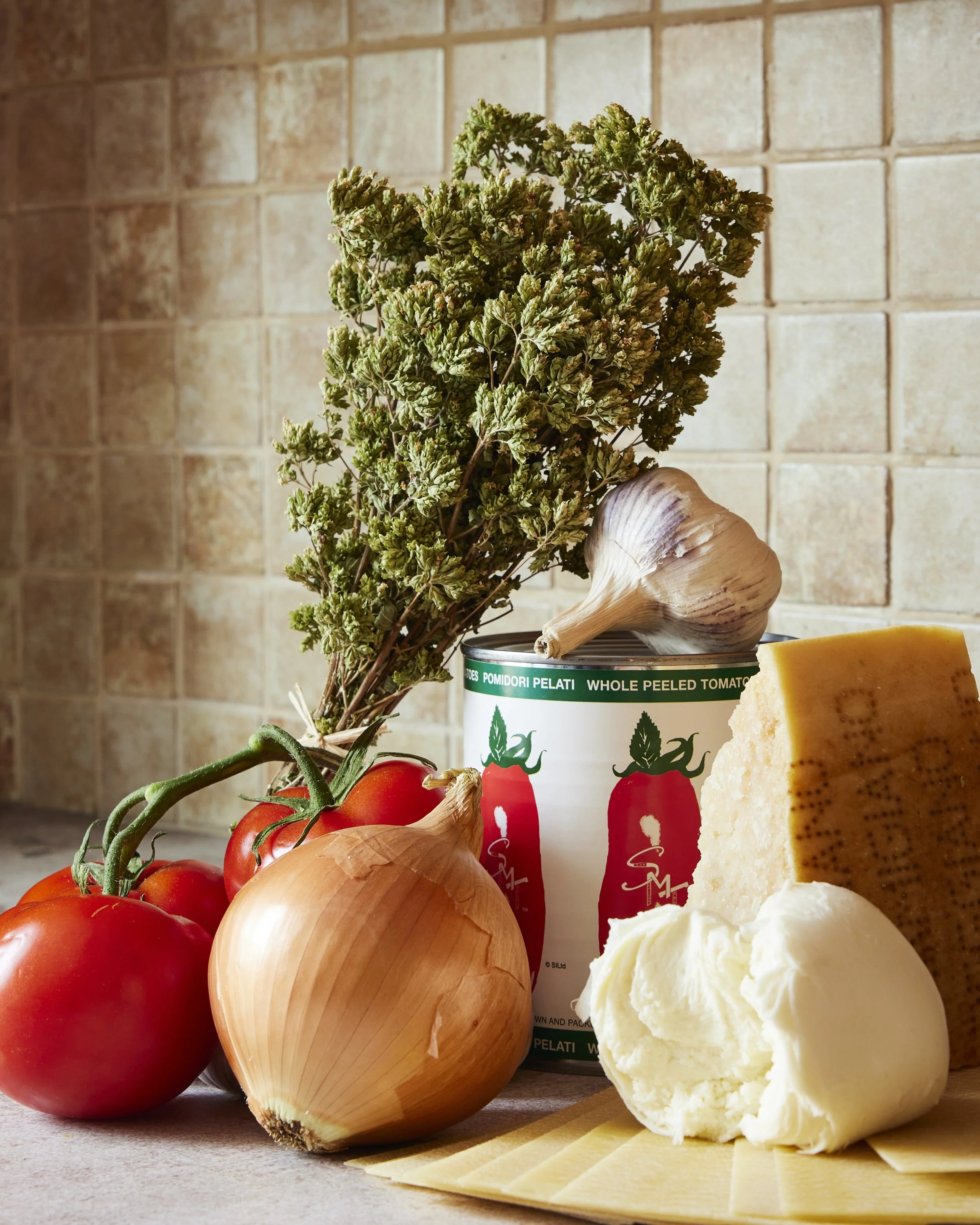Fresh produce including tomatoes, onions, garlic, basil, cheese, and a can of whole peeled tomatoes on a kitchen counter.