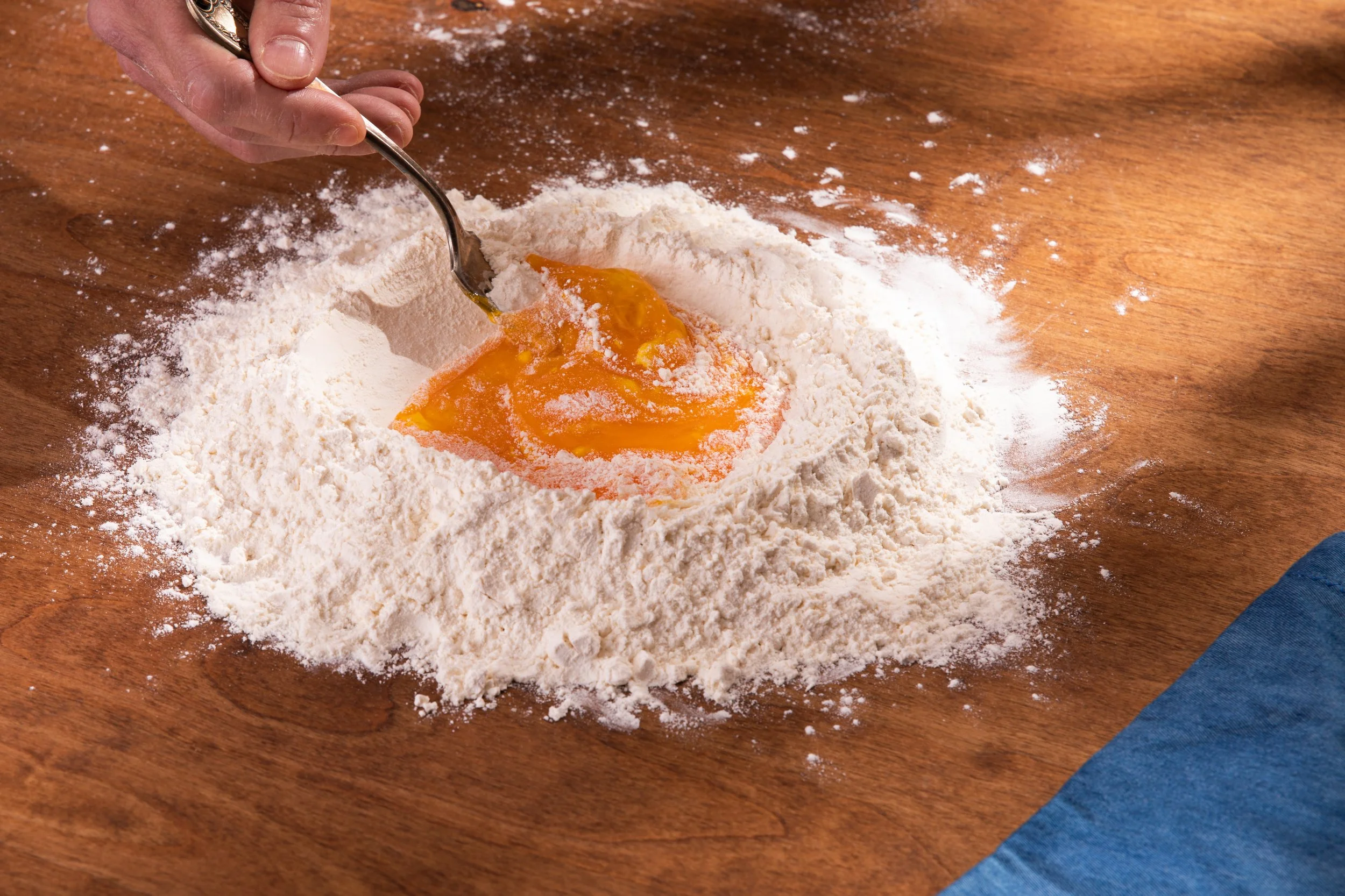 Person mixing egg yolk into a pile of flour on a wooden surface.