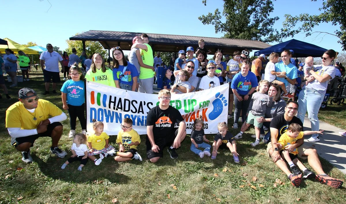 A group of people, including children and adults, gathered outdoors for a walk event supporting Down syndrome awareness. They are holding a large banner that reads "HADSA'S WAK FOR DOWN SYNDROME." Some people are sitting on the grass, and there are tents and trees in the background.