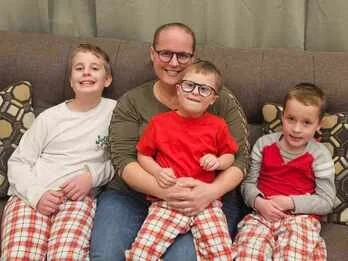 A woman with three young children sitting on a couch, all smiling. The children are wearing plaid pajama pants, and the woman is in glasses and a green shirt.