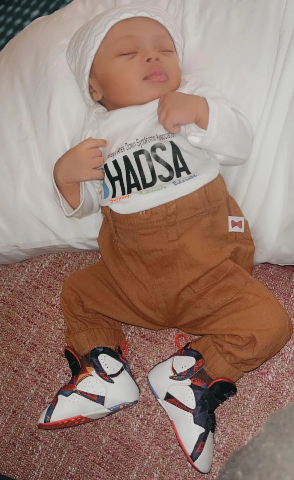 Young baby lying on a bed, wearing a white hat, a shirt with the HADSA logo, brown pants, and colorful sneakers, with eyes closed and fists near the chest.