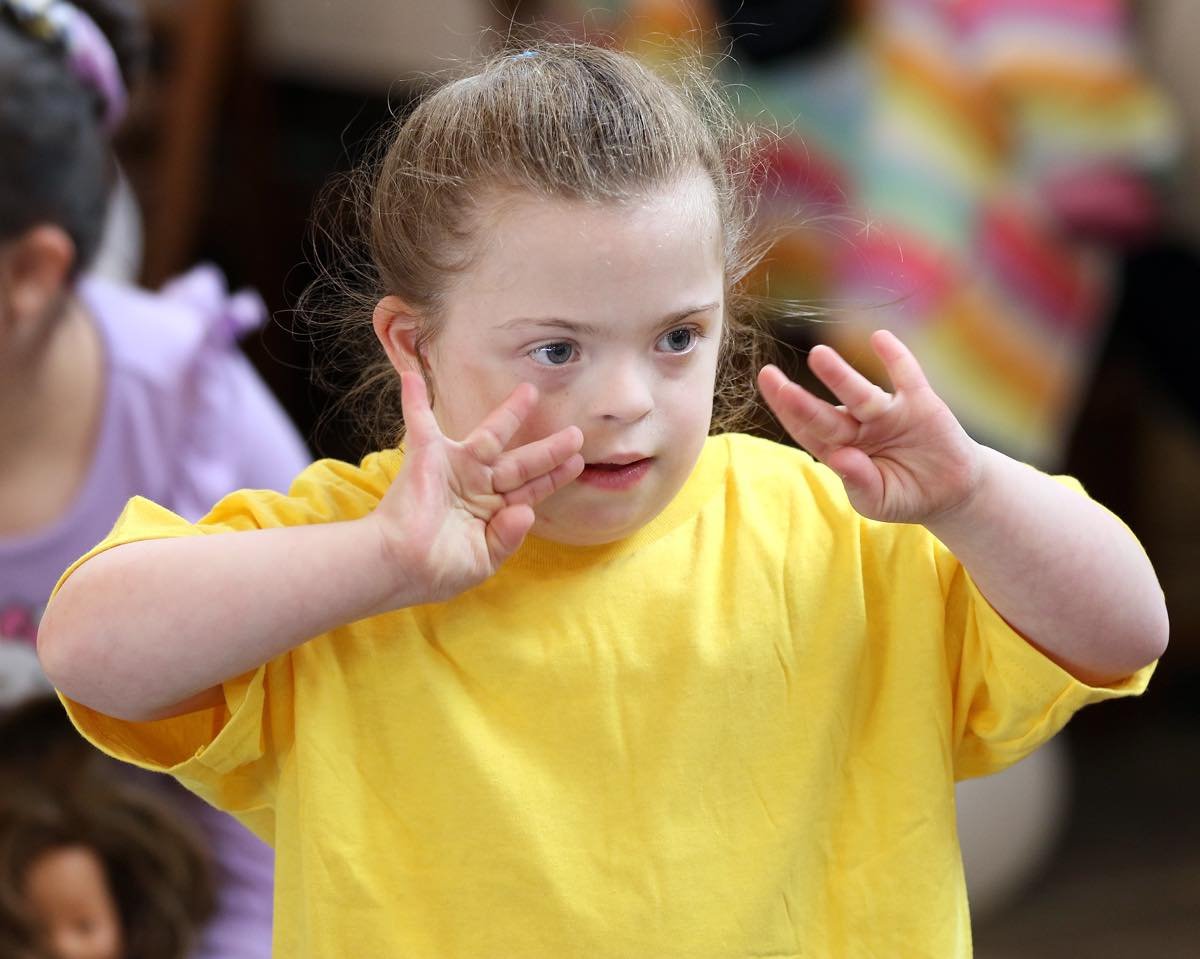 A young girl with red hair in a yellow shirt places her hands near her face in an indoor setting with blurred colorful background.