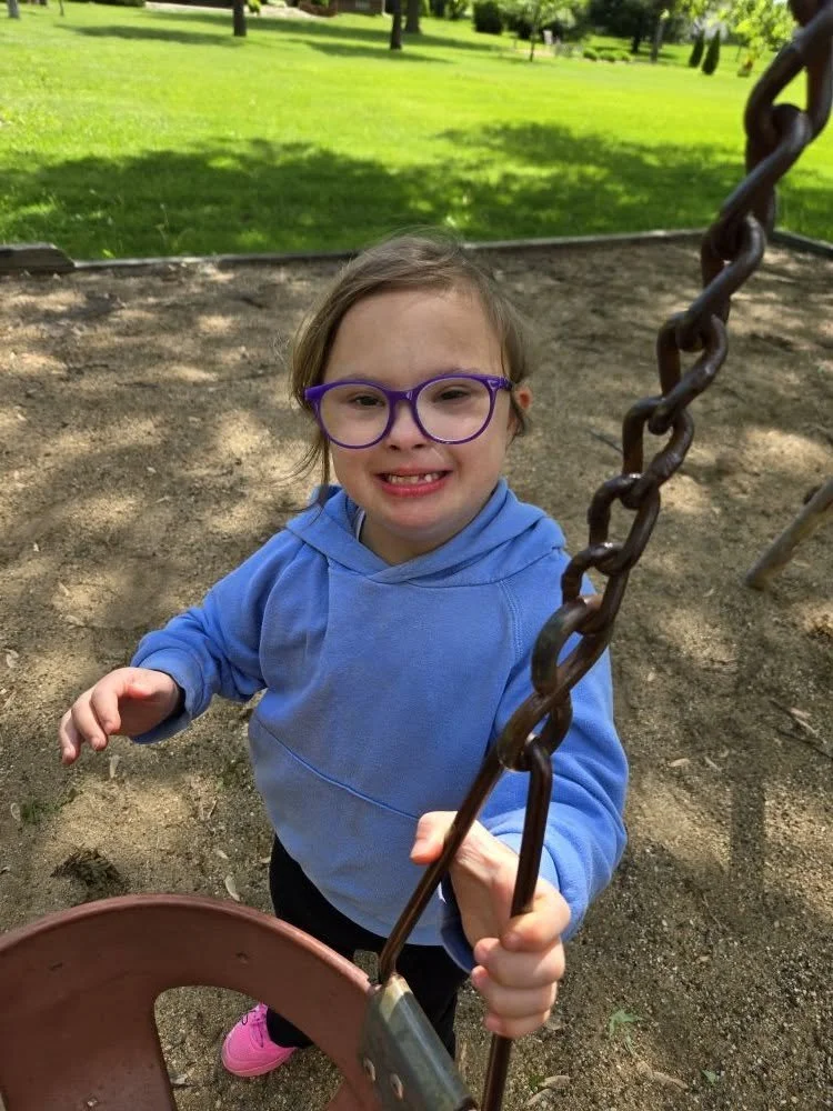 A young girl with glasses and a blue hoodie smiling at the camera while playing on a swing in a park.