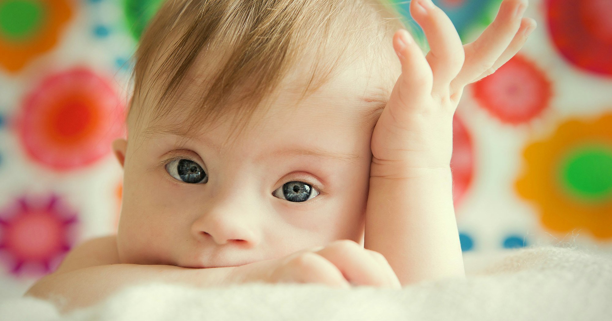 Close-up of a baby with blue eyes resting on a soft surface, with one hand on its forehead and colorful, flower-shaped background.