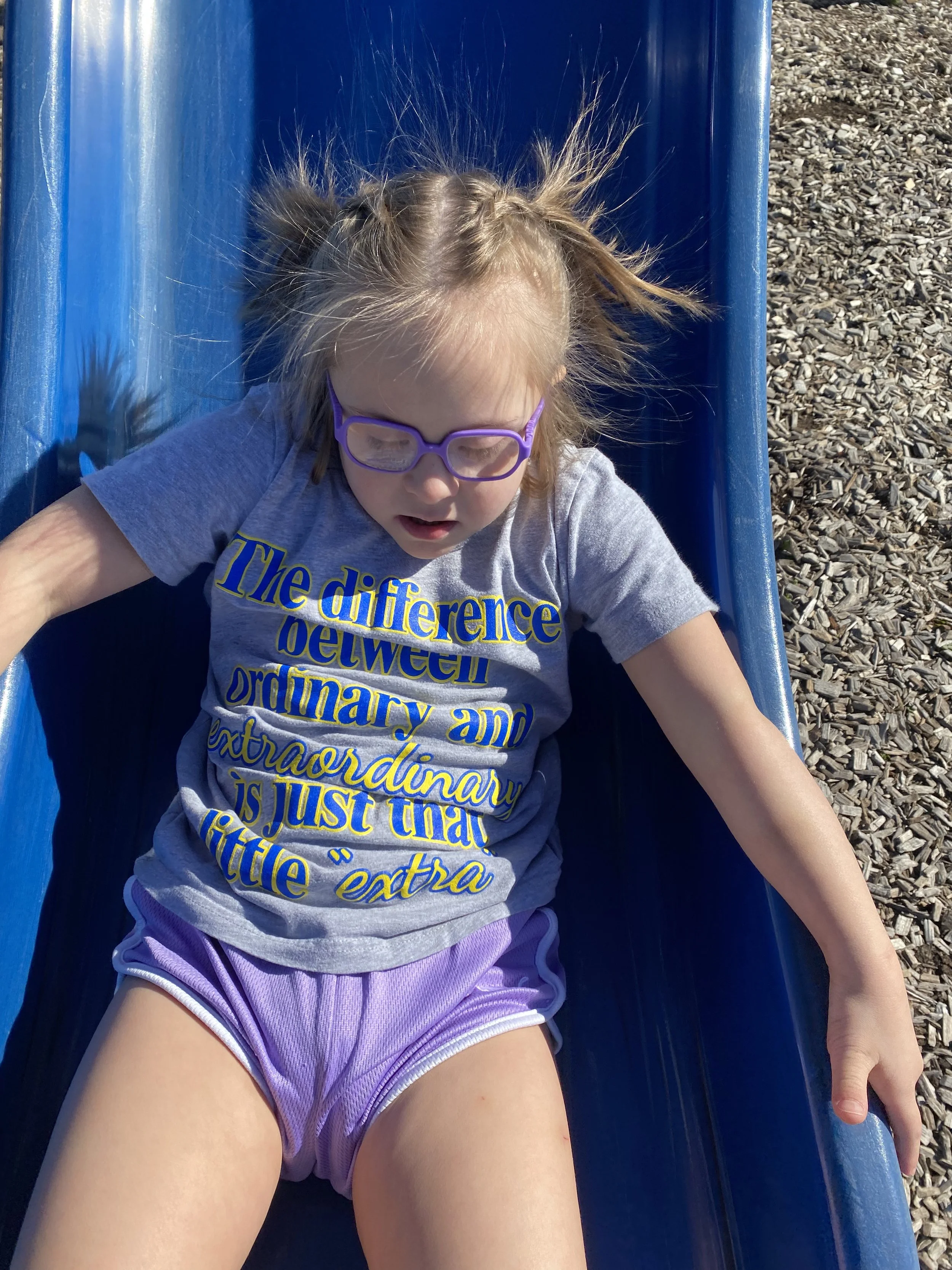 A young girl with glasses, light brown hair tied into pigtails, is sitting at the bottom of a blue slide at a playground. She is wearing a gray T-shirt with yellow and blue text and pink shorts. The girl appears to be tired or resting after sliding.