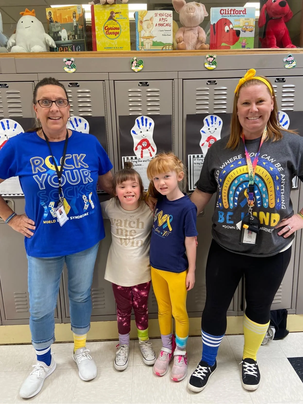 Two women and two young girls standing in front of lockers decorated with colorful handprint posters, in a school hallway or classroom.