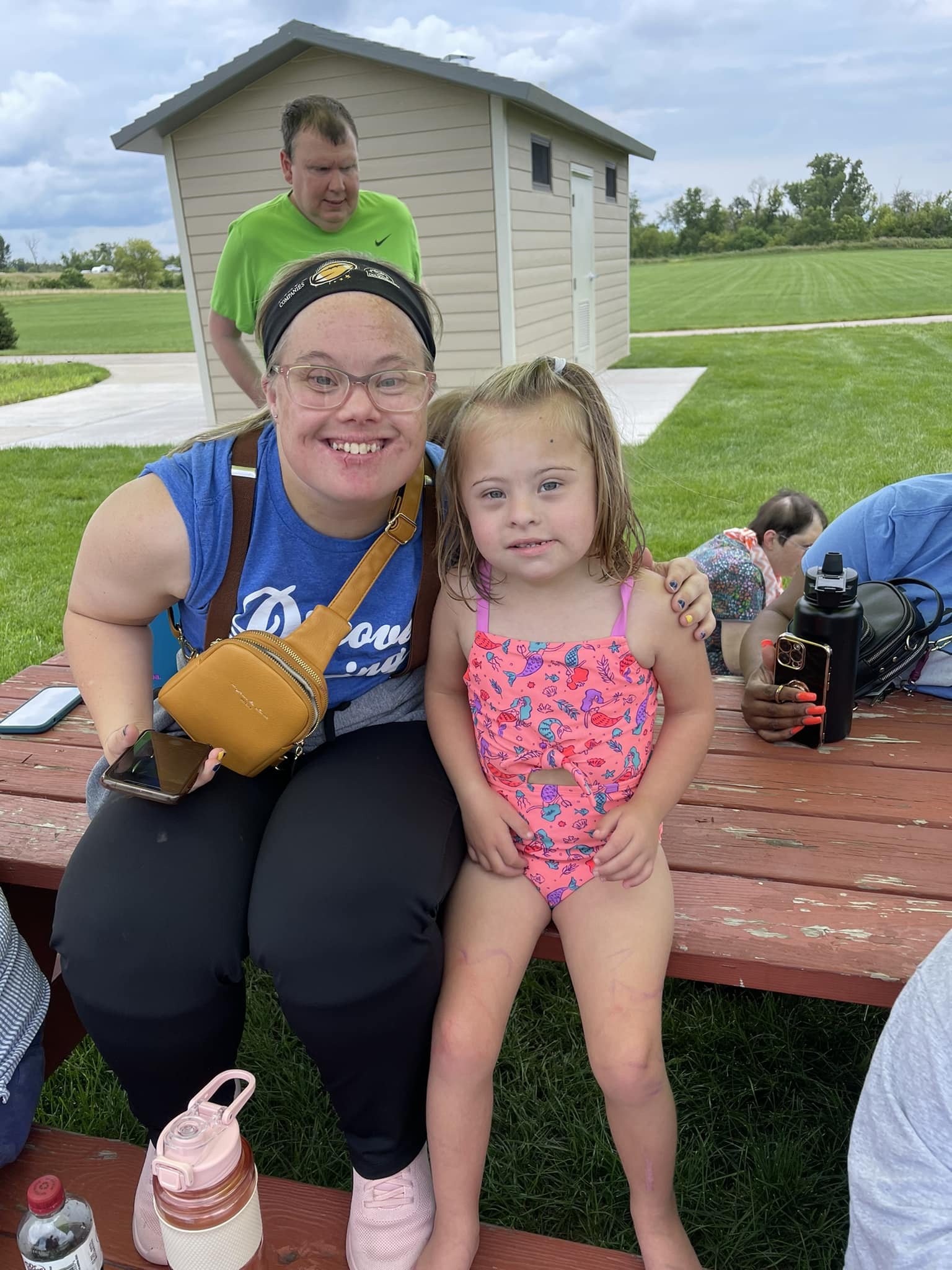 A smiling woman with glasses and a blue shirt sitting on a bench next to a young girl in a pink swimsuit, posing for a photo outdoors with a man in a green shirt in the background and several people around them.