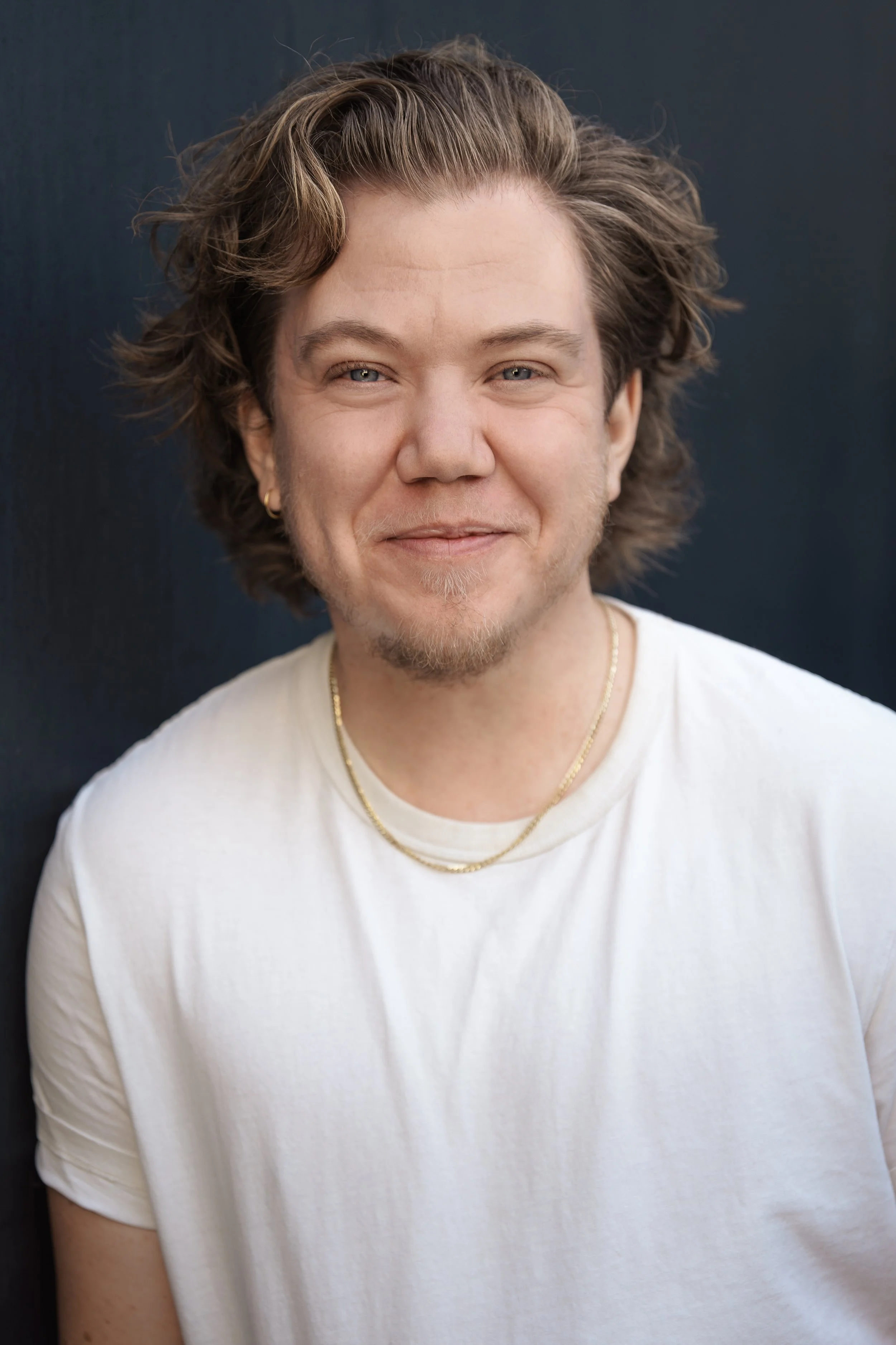 A young man with wavy brown hair, light blue eyes, and a light beard, smiling in front of a dark background, wearing a white t-shirt and gold necklaces.