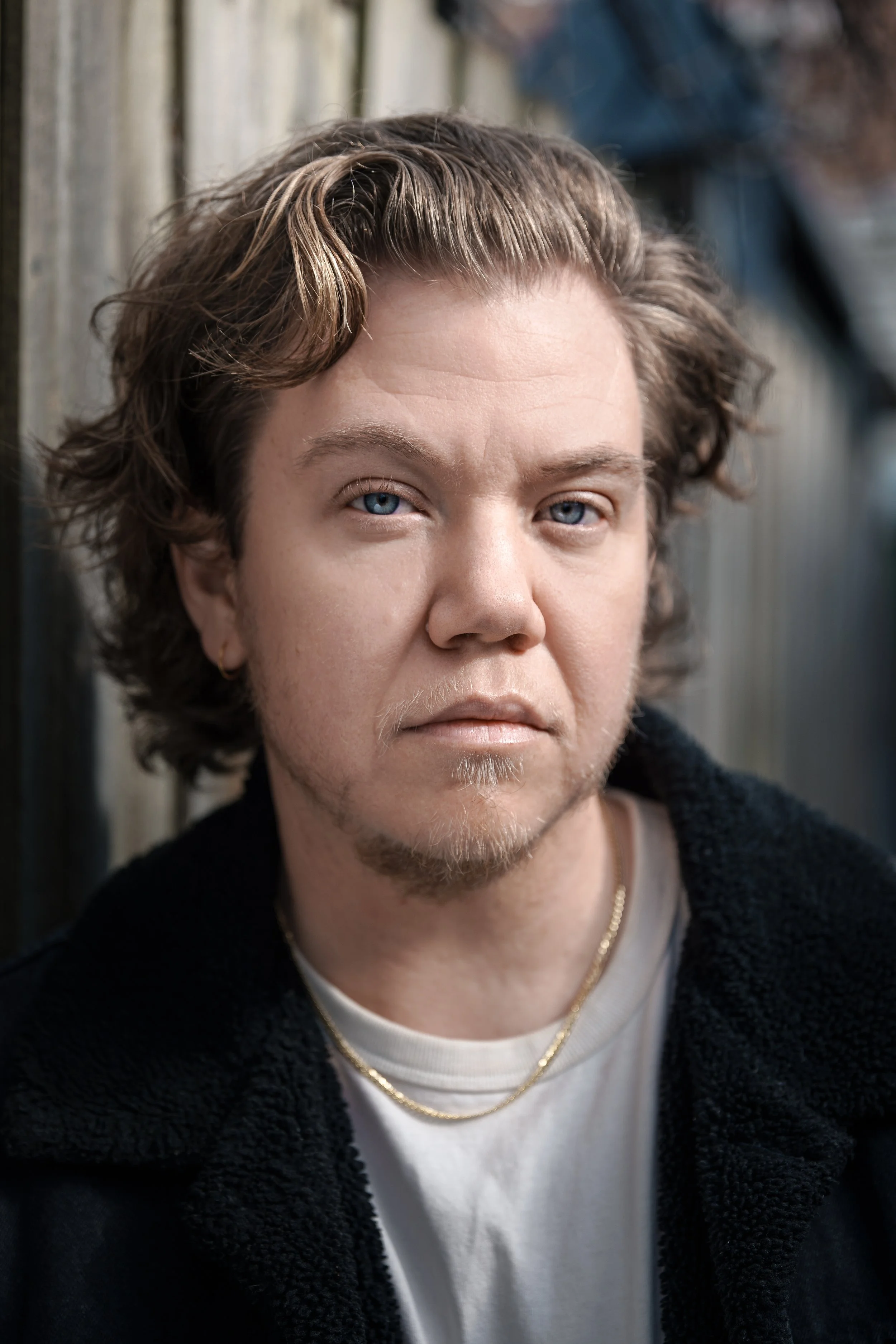 Portrait of a young man with wavy brown hair, blue eyes, wearing a black jacket, gold chain necklace, and a white shirt, standing outdoors with a wooden fence in the background.