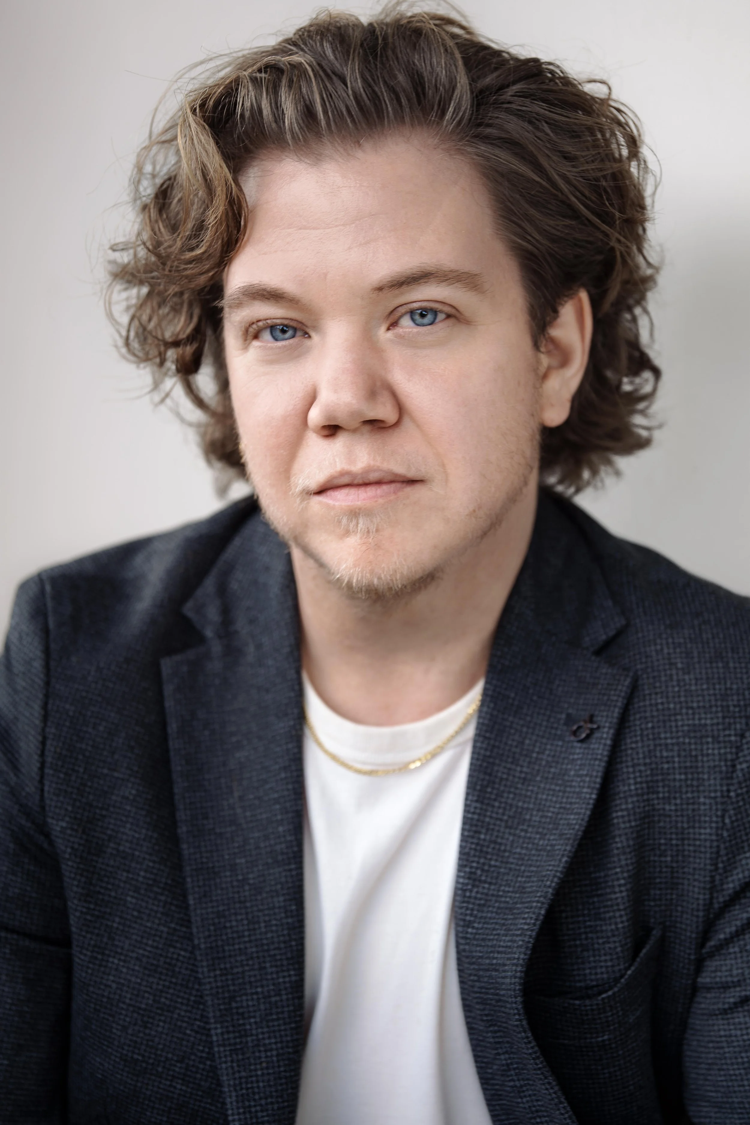 Portrait of a young man with shoulder-length wavy brown hair and blue eyes, wearing a dark blazer over a white shirt and gold necklaces, against a plain light background.