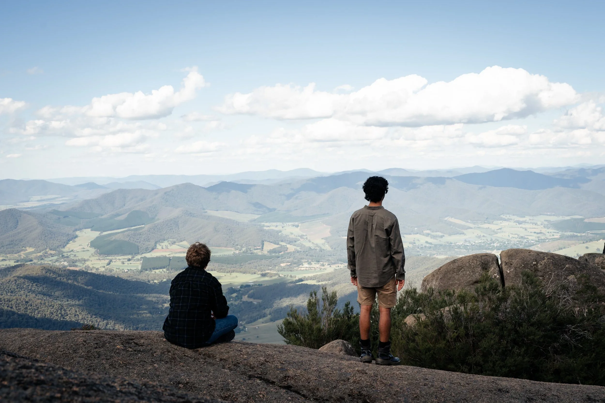 Two people are on a rocky hillside overlooking a valley with rolling hills and mountains under a partly cloudy sky.