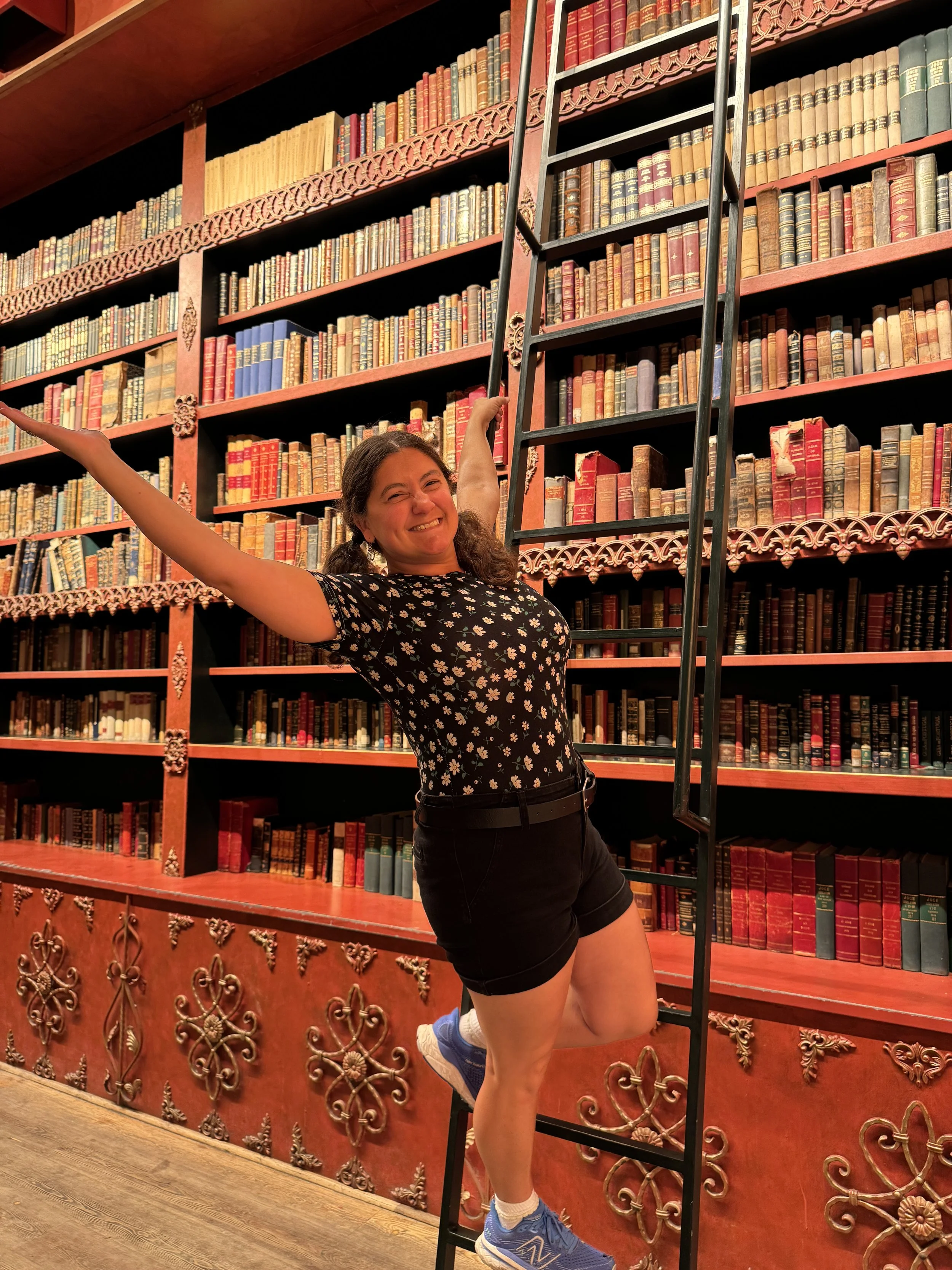 A woman with dark hair in pigtails standing on a ladder in front of a large bookshelf filled with colorful old books, smiling with her arms stretched out like Belle from Disney's Beauty and the Beast. Every readers dream.