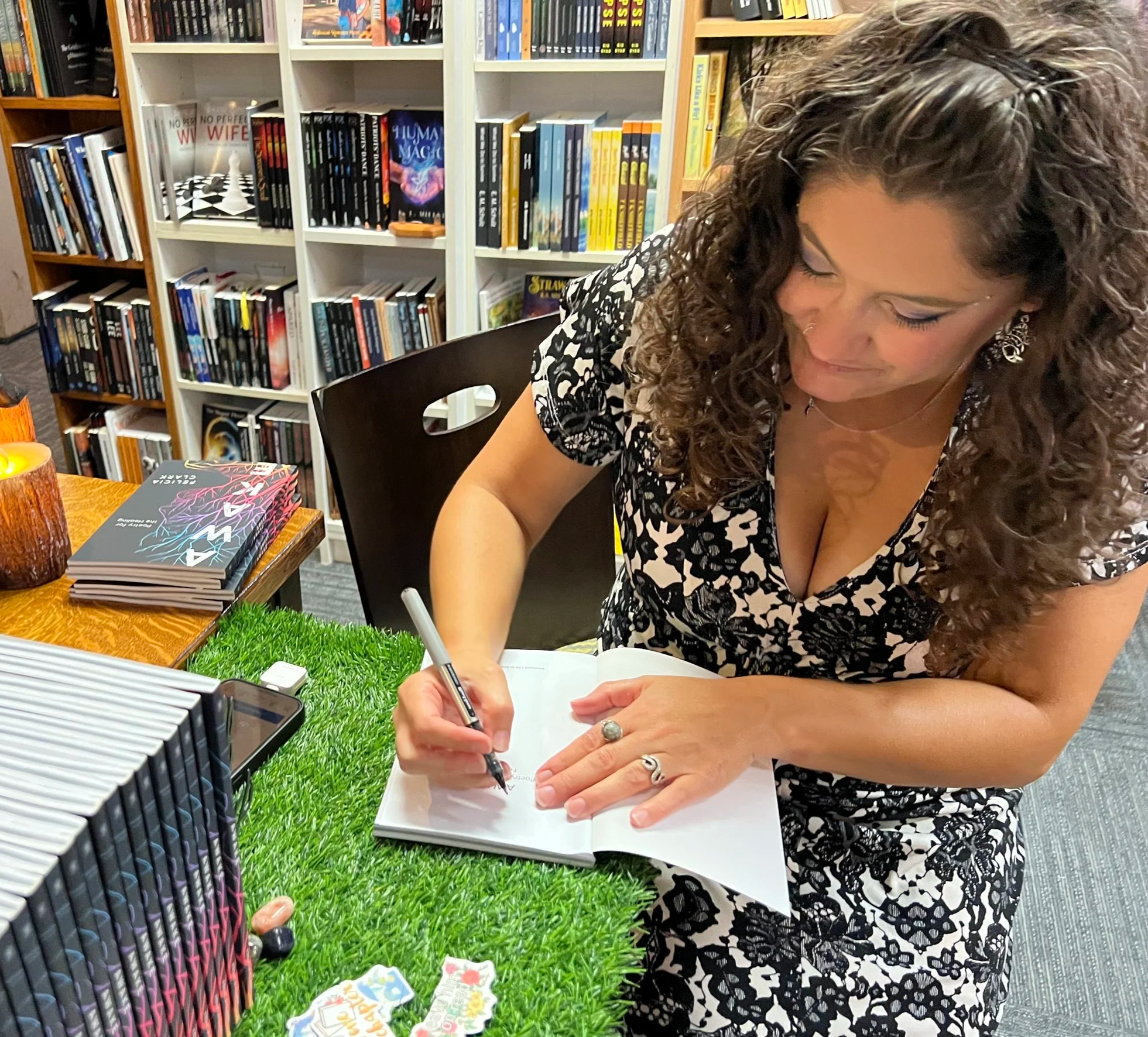 A woman author with curly hair in a black and white lace dress is signing her published book at a table in a Wisconsin bookstore.