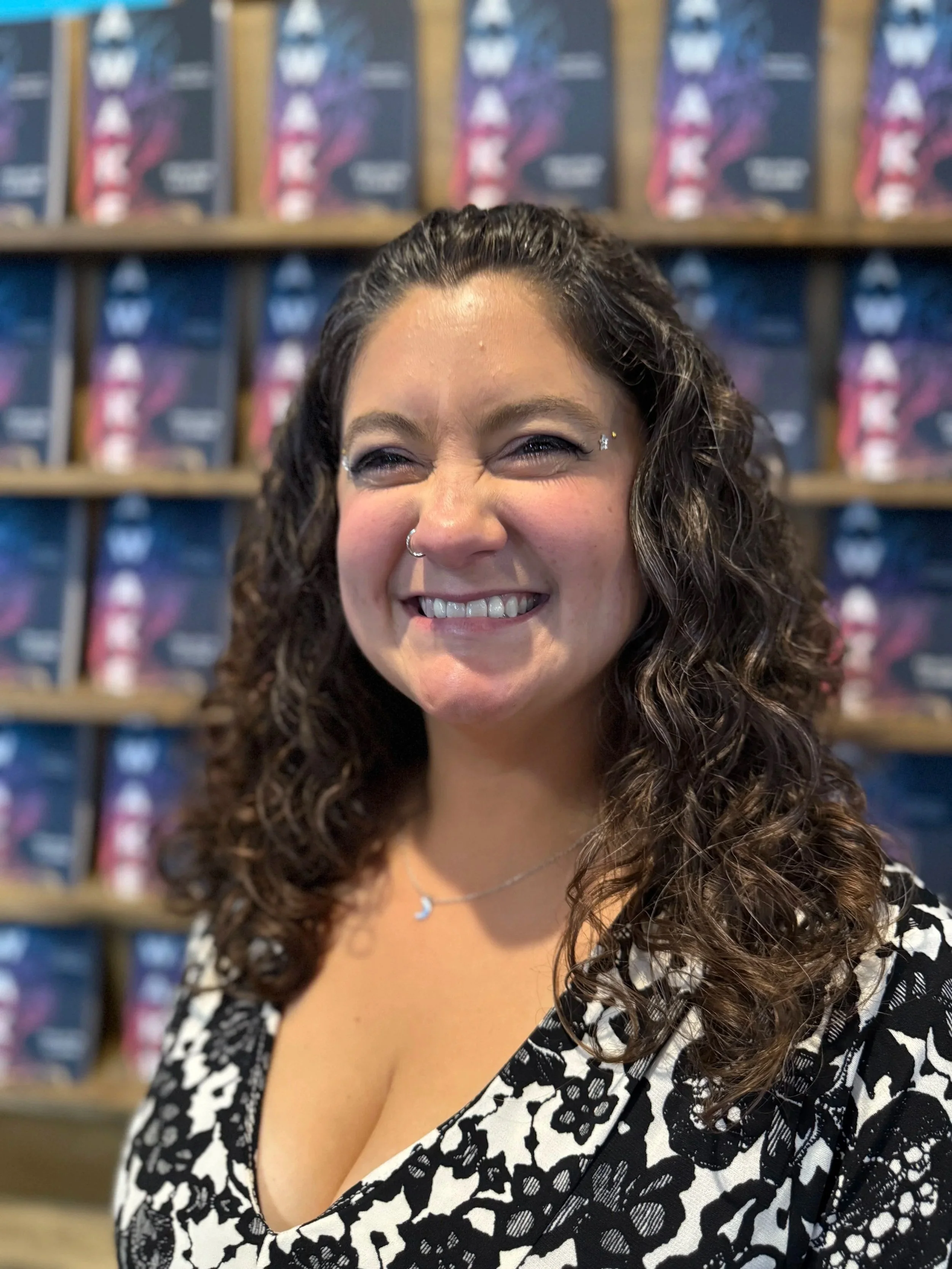 A woman author with curly brown hair smiling at the camera, wearing a black and white dress, standing in front of bookshelves filled with her published books at her book launch event in Wisconsin.