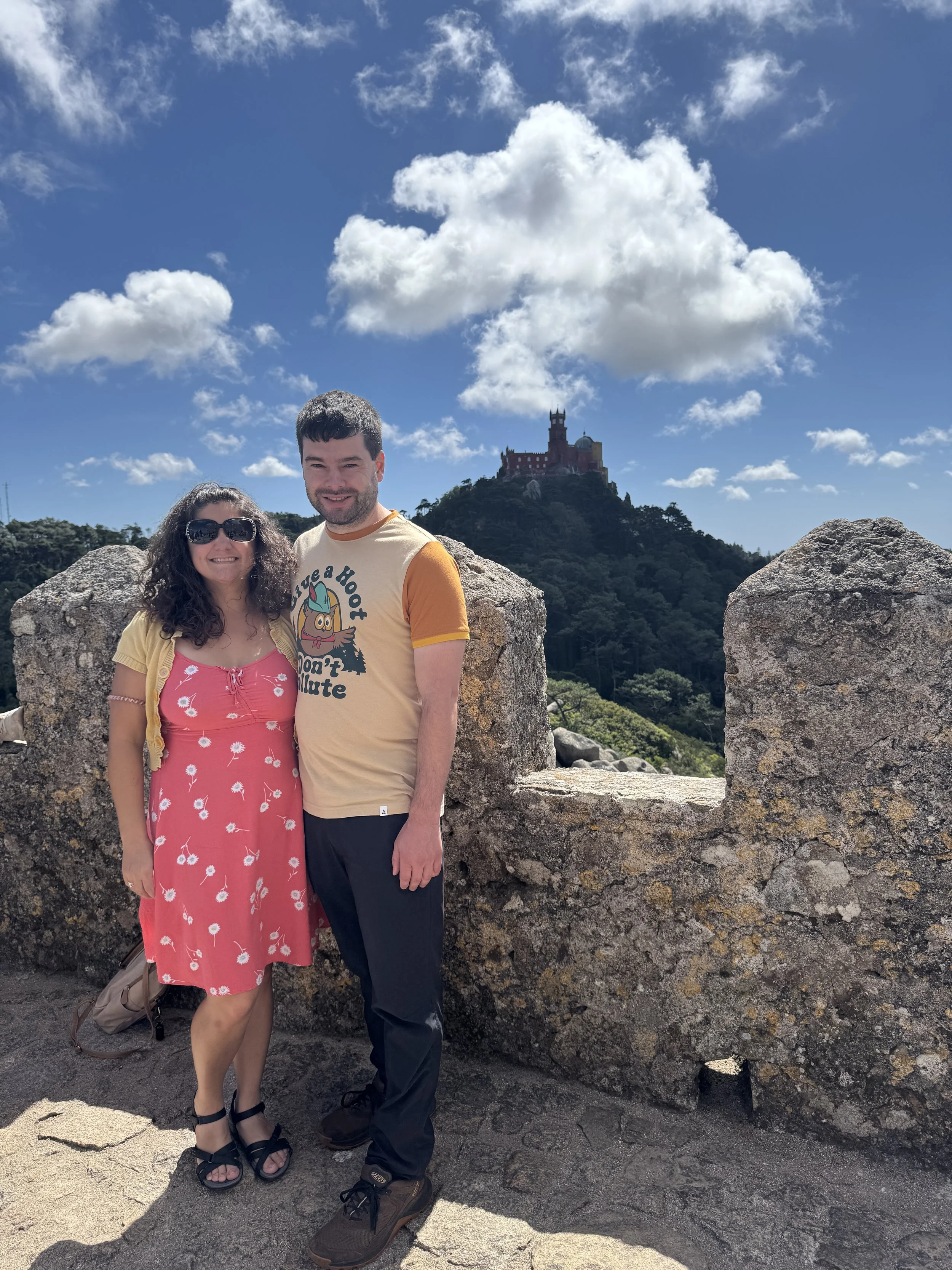 Bookstore owners and building owner stand in front of a stone wall with a castle on a hill in the background under a partly cloudy sky during their visit to Portugal.