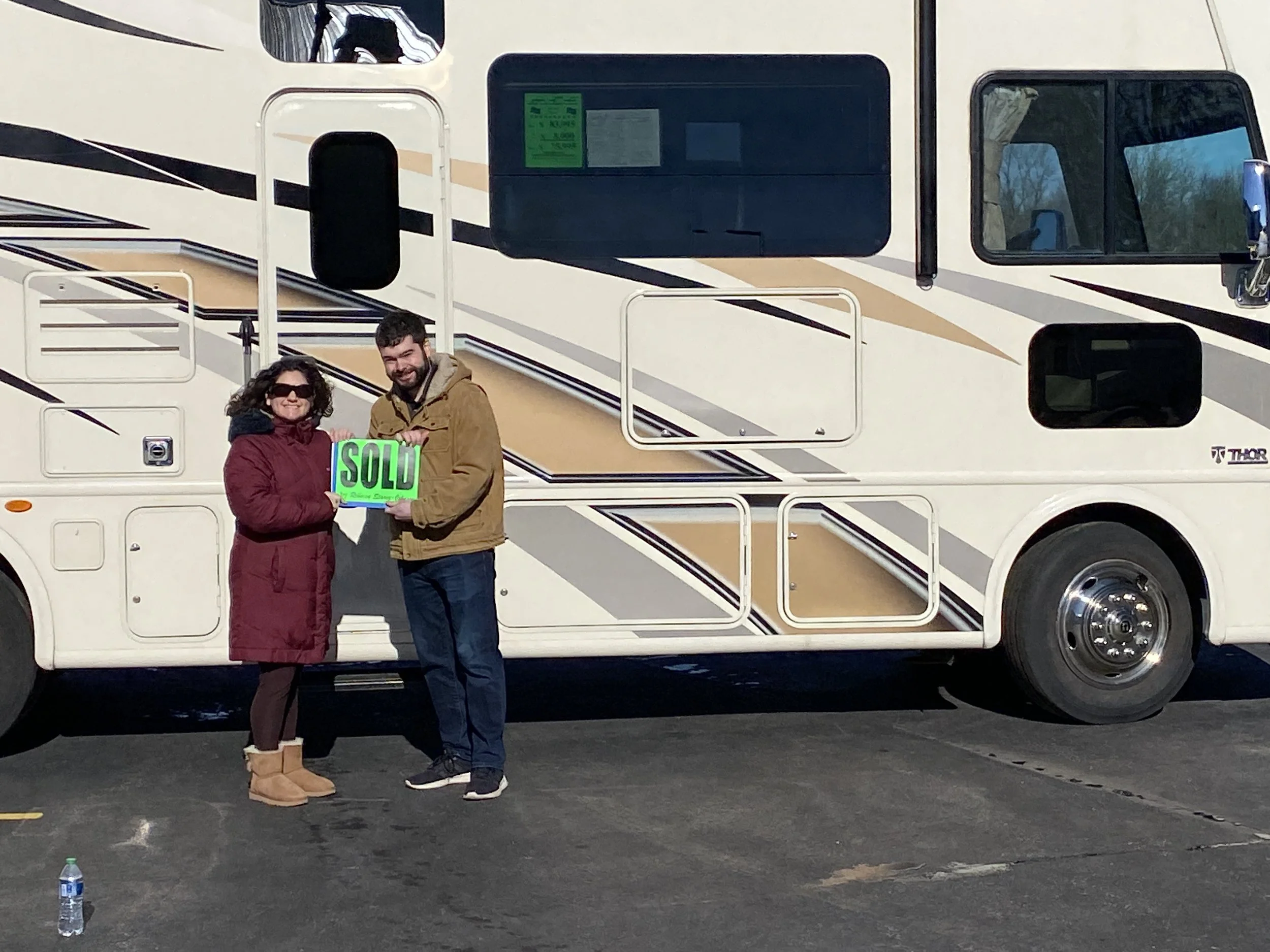 A woman and a man standing in front of their RV, smiling and holding a green sign that says 'SOLD.' They are about to embark on a nomad adventure throughout the United States of America.