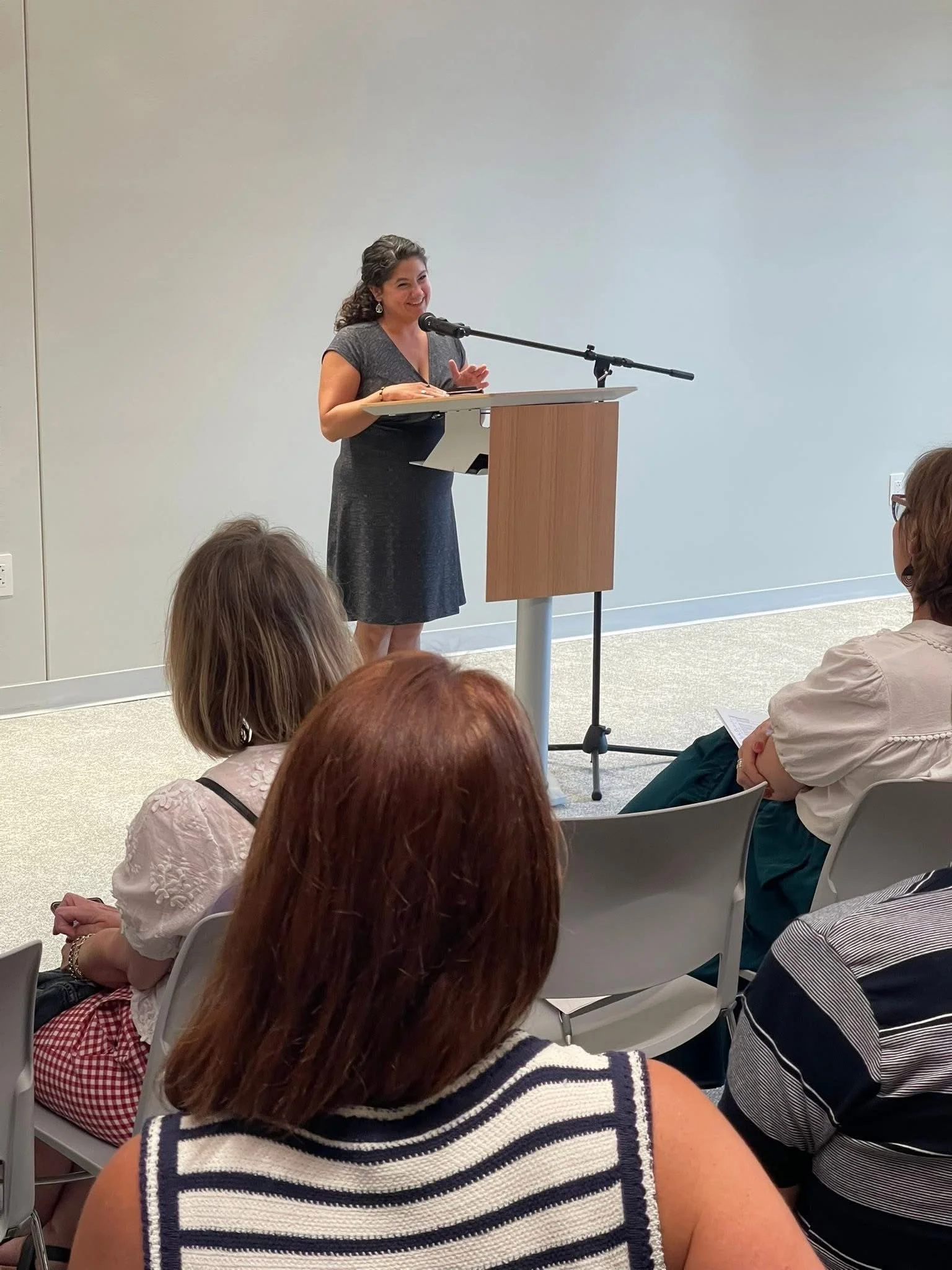 A poet and author in a gray dress reads from her poetry memoir book at a podium with a microphone in front of an audience in the Appleton Public Library in Wisconsin for a read your favorite poem event.
