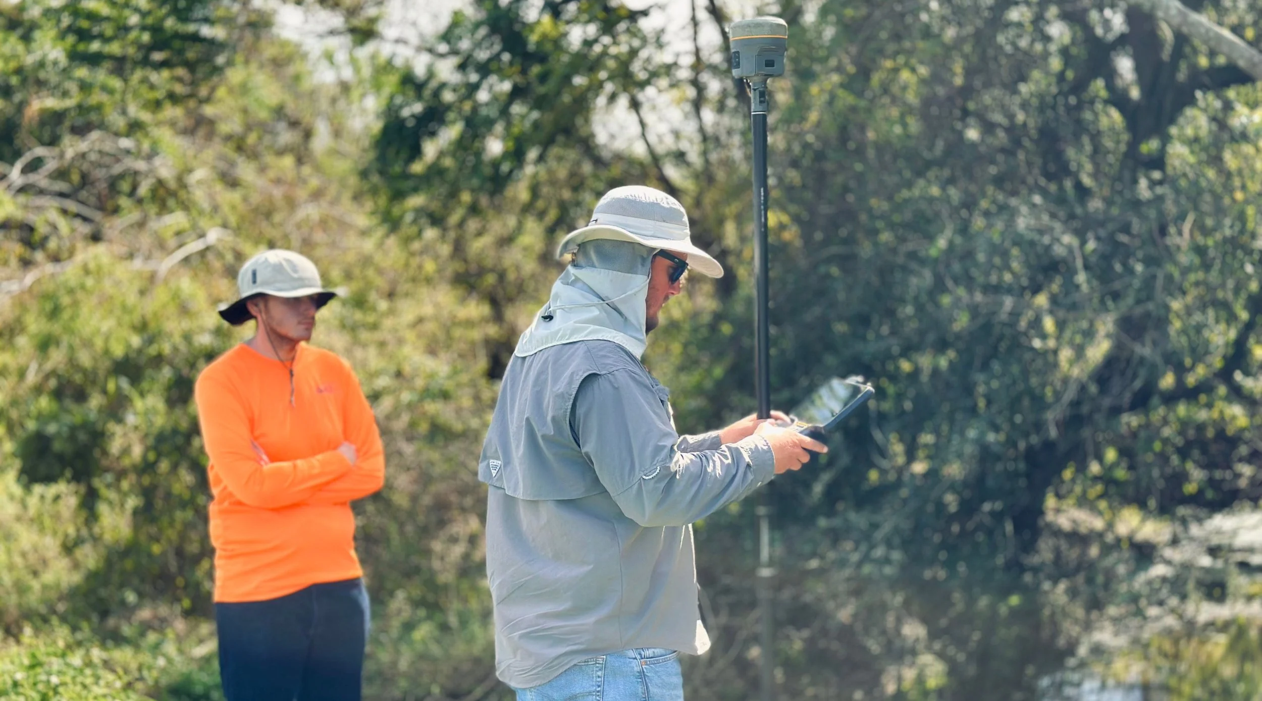 Two individuals outdoors with trees in the background, one using a tablet and the other observing, both wearing wide-brimmed hats and light outdoor clothing.