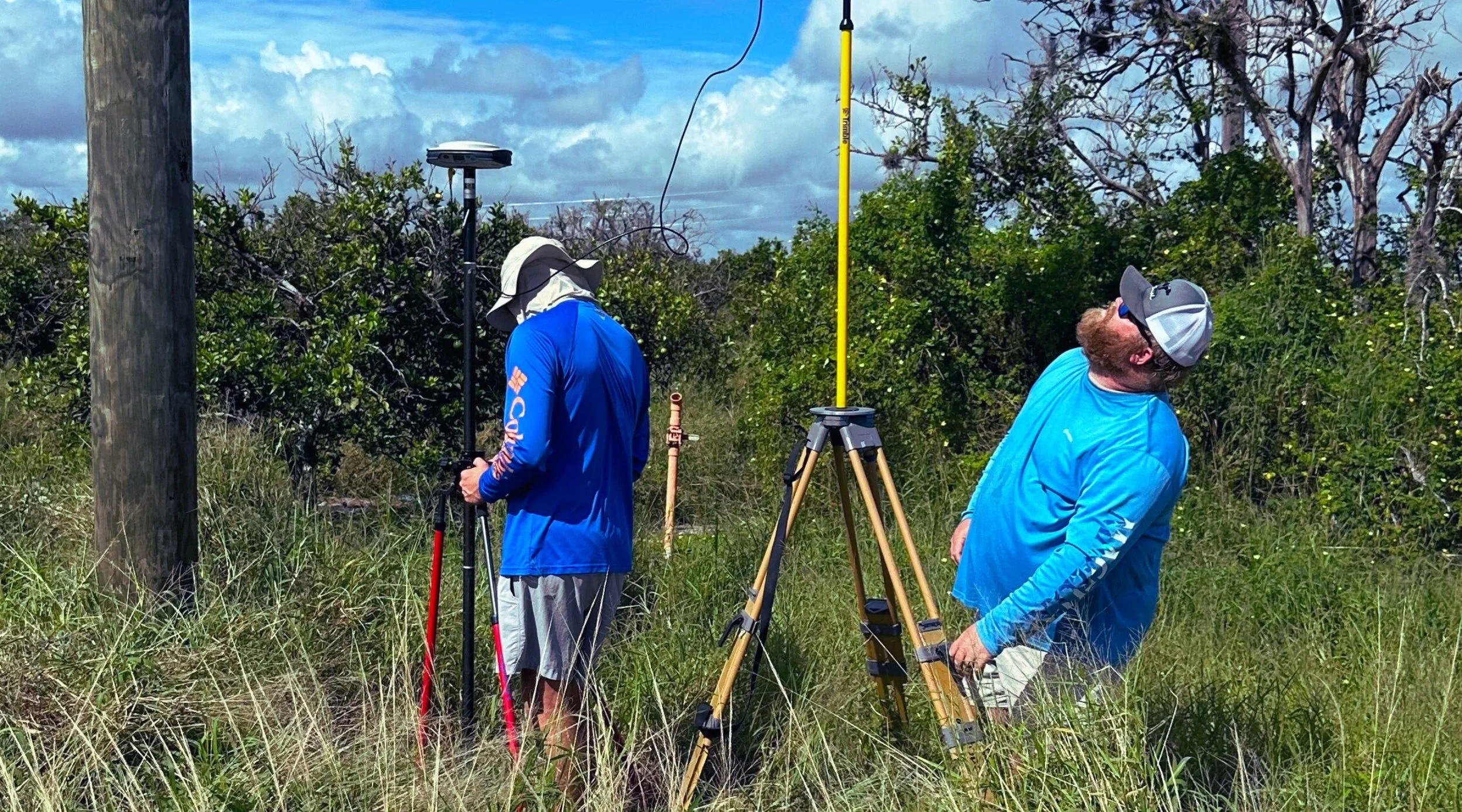 Two men in blue shirts conducting land surveying work in a grassy outdoor area, with surveying equipment including a tripod and a measuring rod, against a backdrop of trees and a partly cloudy sky.