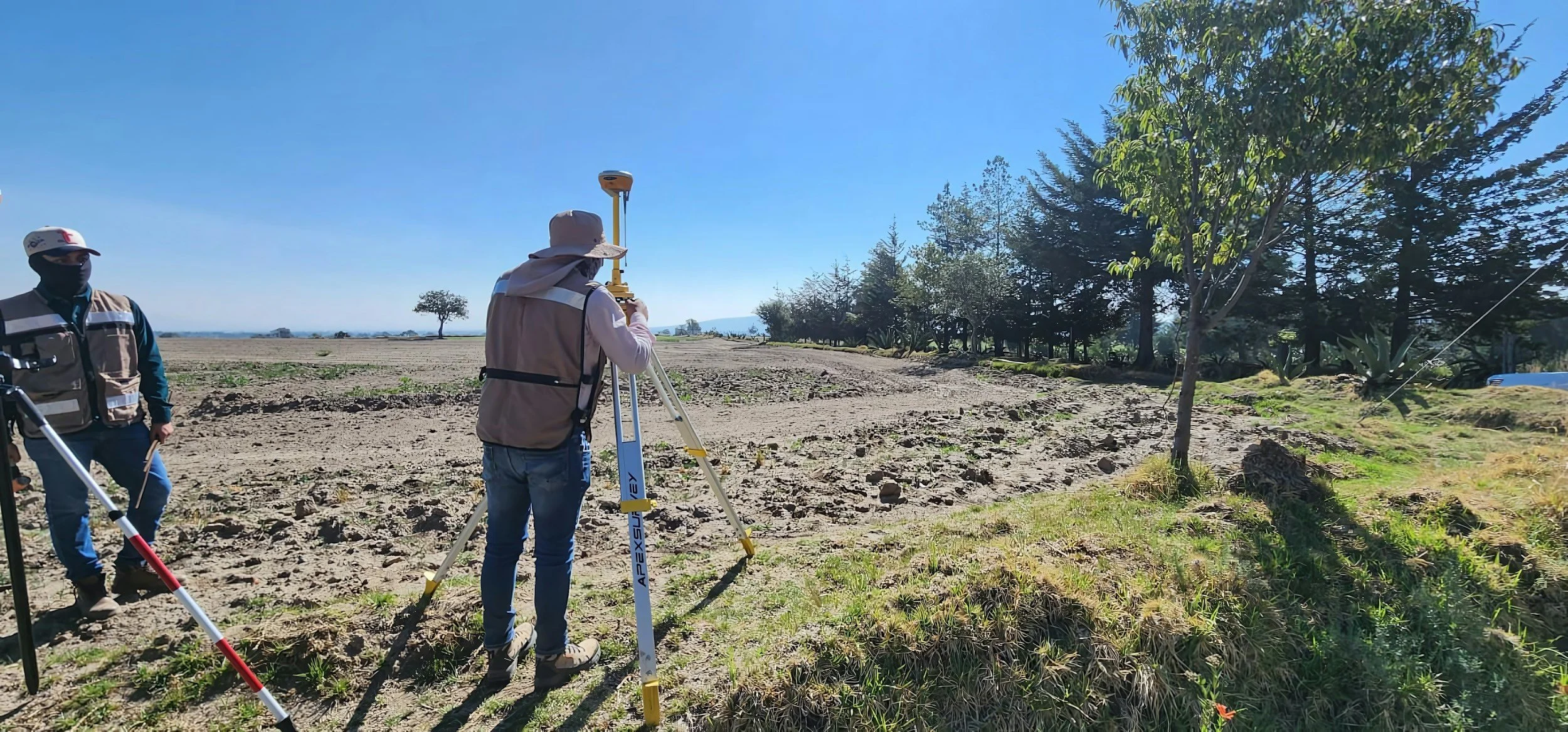 Two surveyors are working in an open field under a clear blue sky. One person is using a total station instrument, and the other is standing nearby with a measuring rod, both wearing vests and hats.