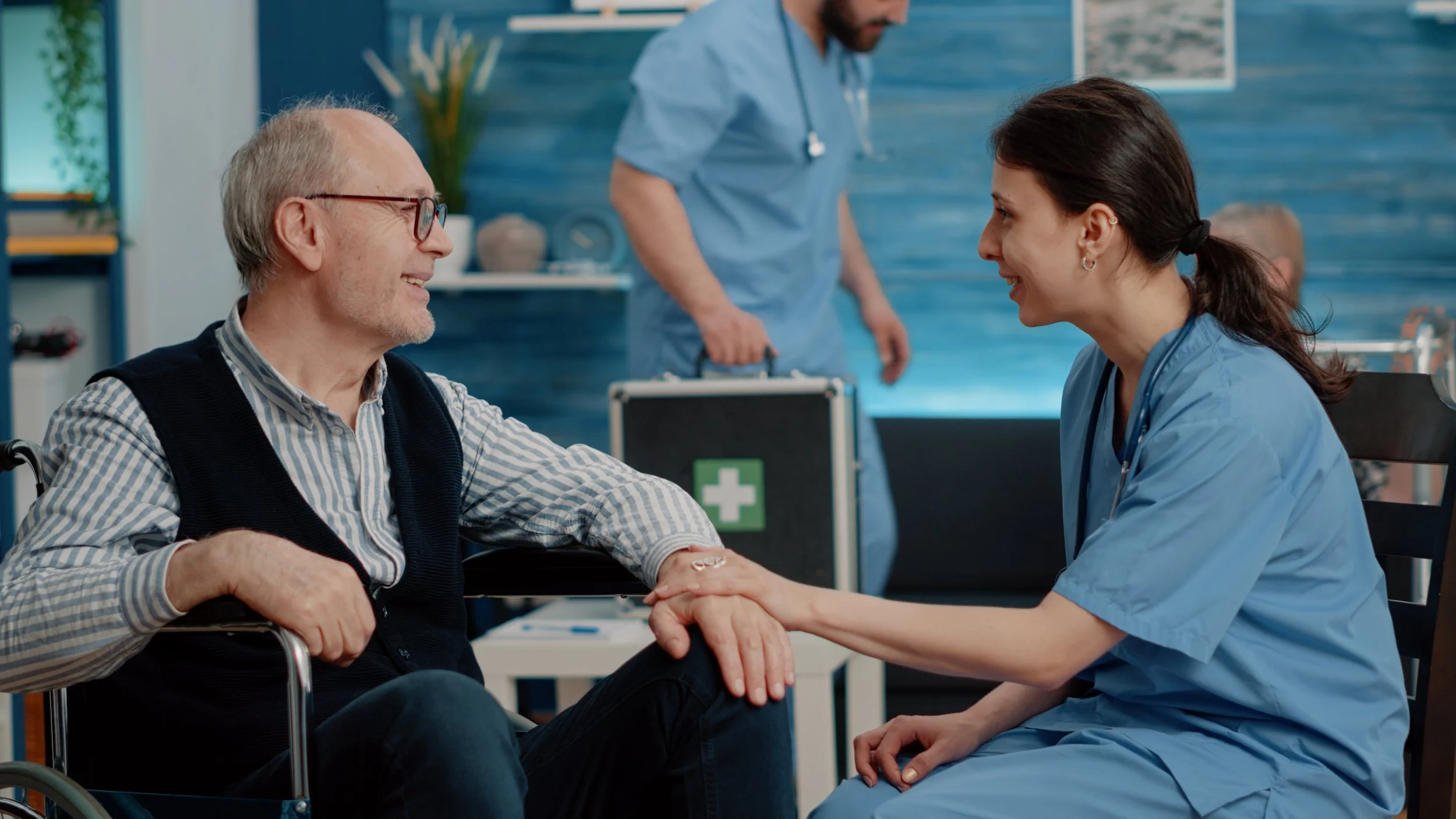 A healthcare worker and an elderly man smiling and holding hands in a medical setting.