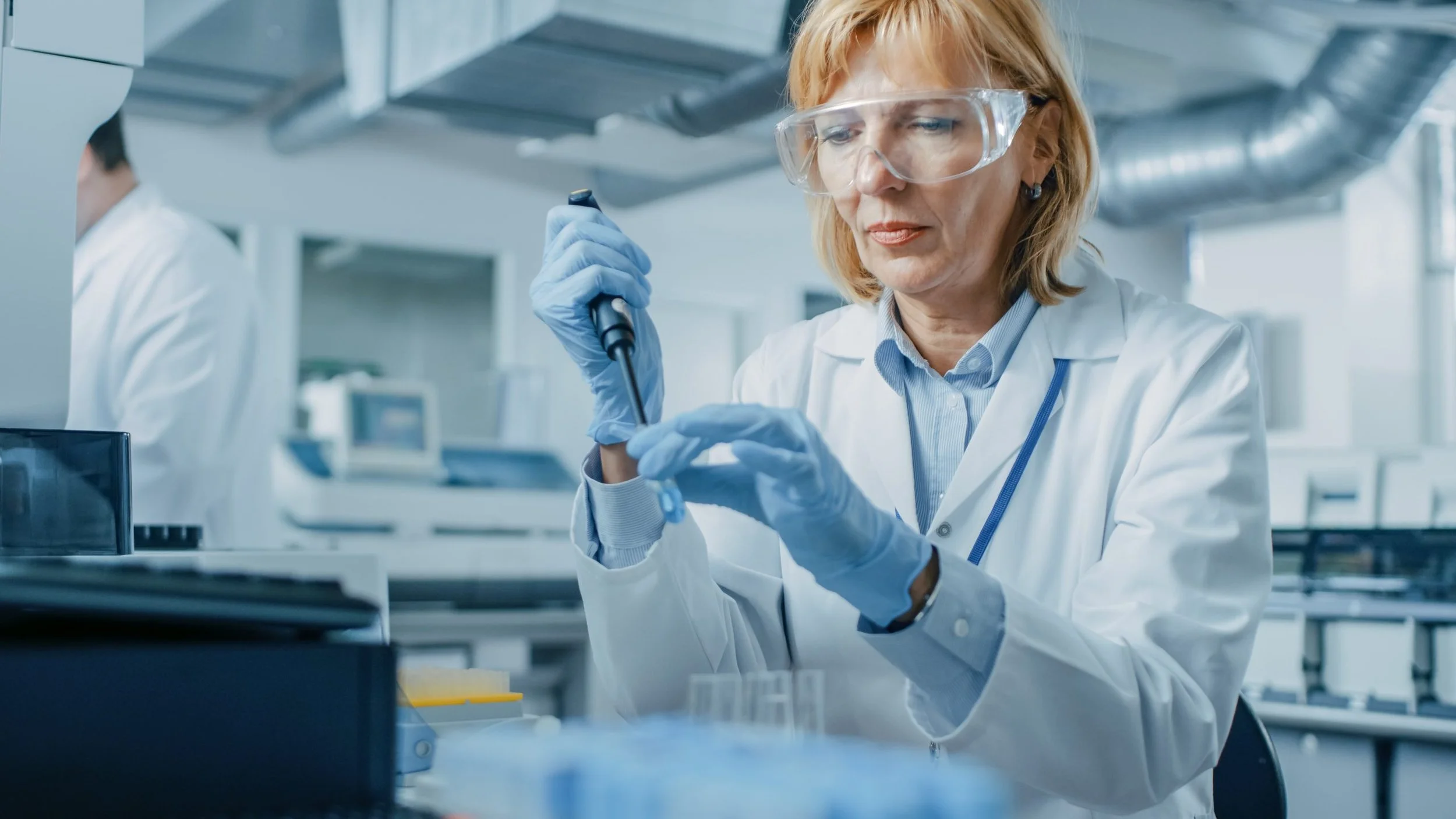 A female scientist wearing safety goggles and blue gloves working with a pipette in a laboratory.