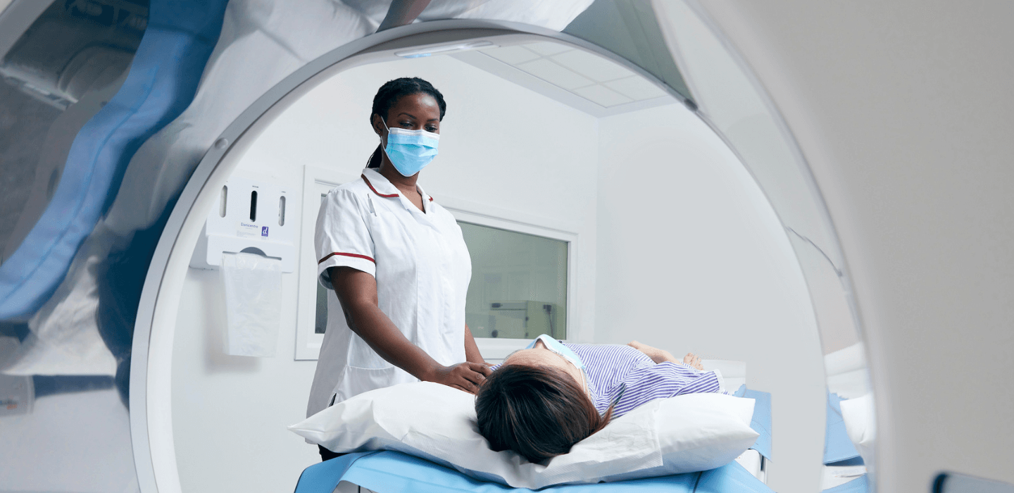 A nurse wearing a face mask and white uniform stands beside a patient lying on a hospital bed, preparing to perform a medical scan inside a large medical imaging machine.