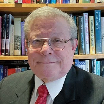 A man with glasses and gray hair wearing a suit and red tie, standing in front of a bookshelf filled with books.