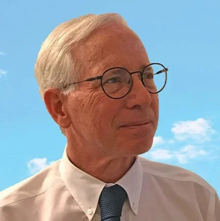 An older man with white hair and glasses, wearing a white shirt and blue tie, against a blue sky with some clouds.