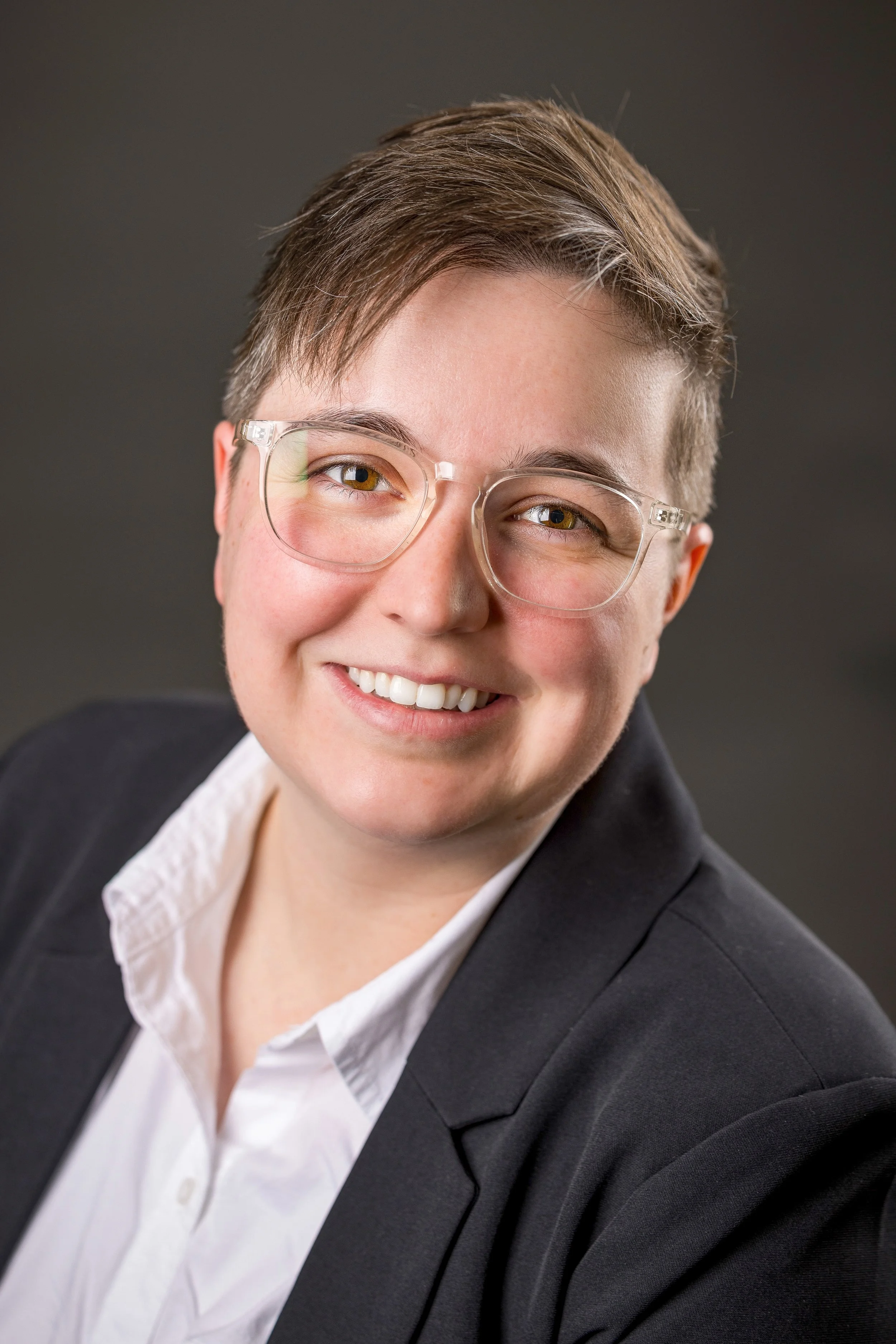 Professional headshot of a smiling woman with short brown hair, glasses, and a black blazer over a white shirt, against a dark gray background.