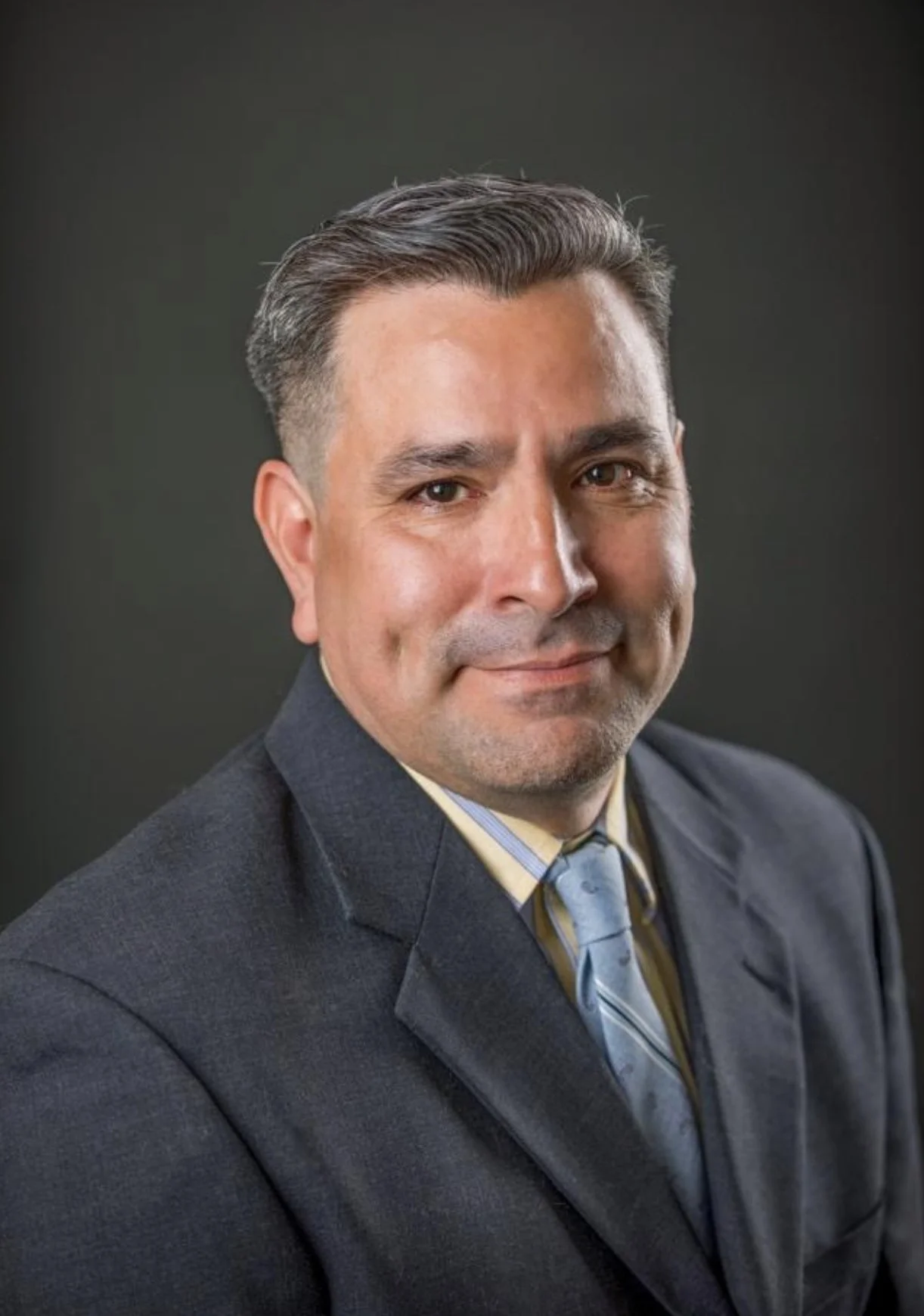 A professional headshot of a man wearing a dark suit, light shirt, and striped tie, smiling slightly against a dark background.