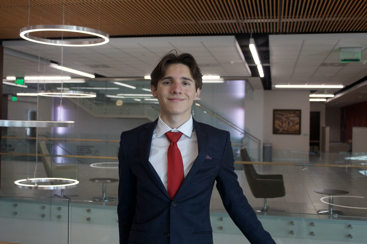Young man in a dark suit and red tie smiling in a modern office lobby.