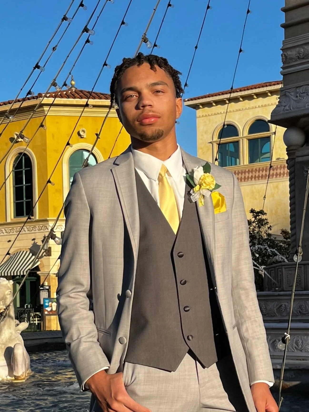 A young man in formal attire standing outdoors on a sunny day with a building and fountain in the background.