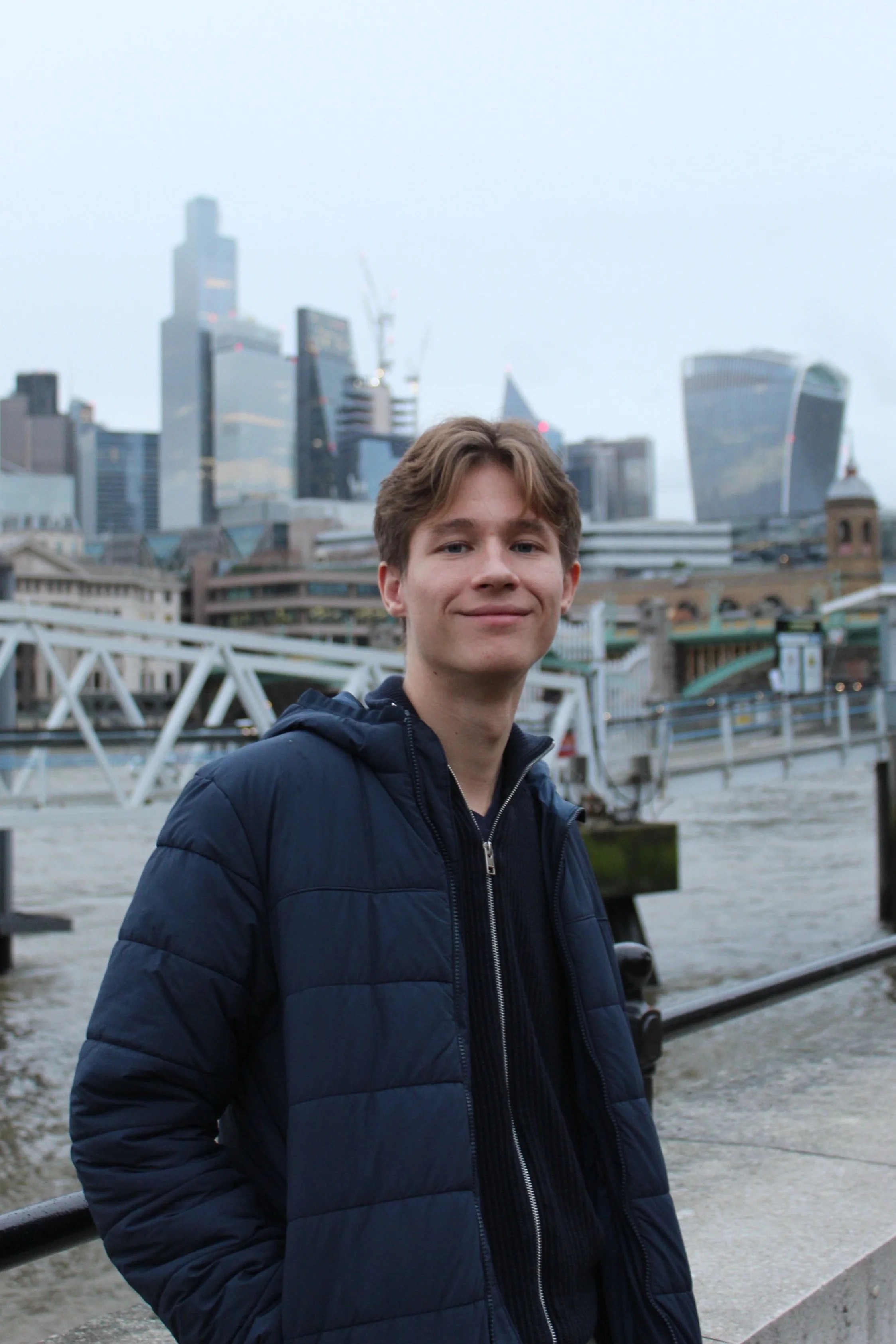 A young man in a navy puffer jacket standing outdoors with a city skyline of tall skyscrapers behind him, near a river.