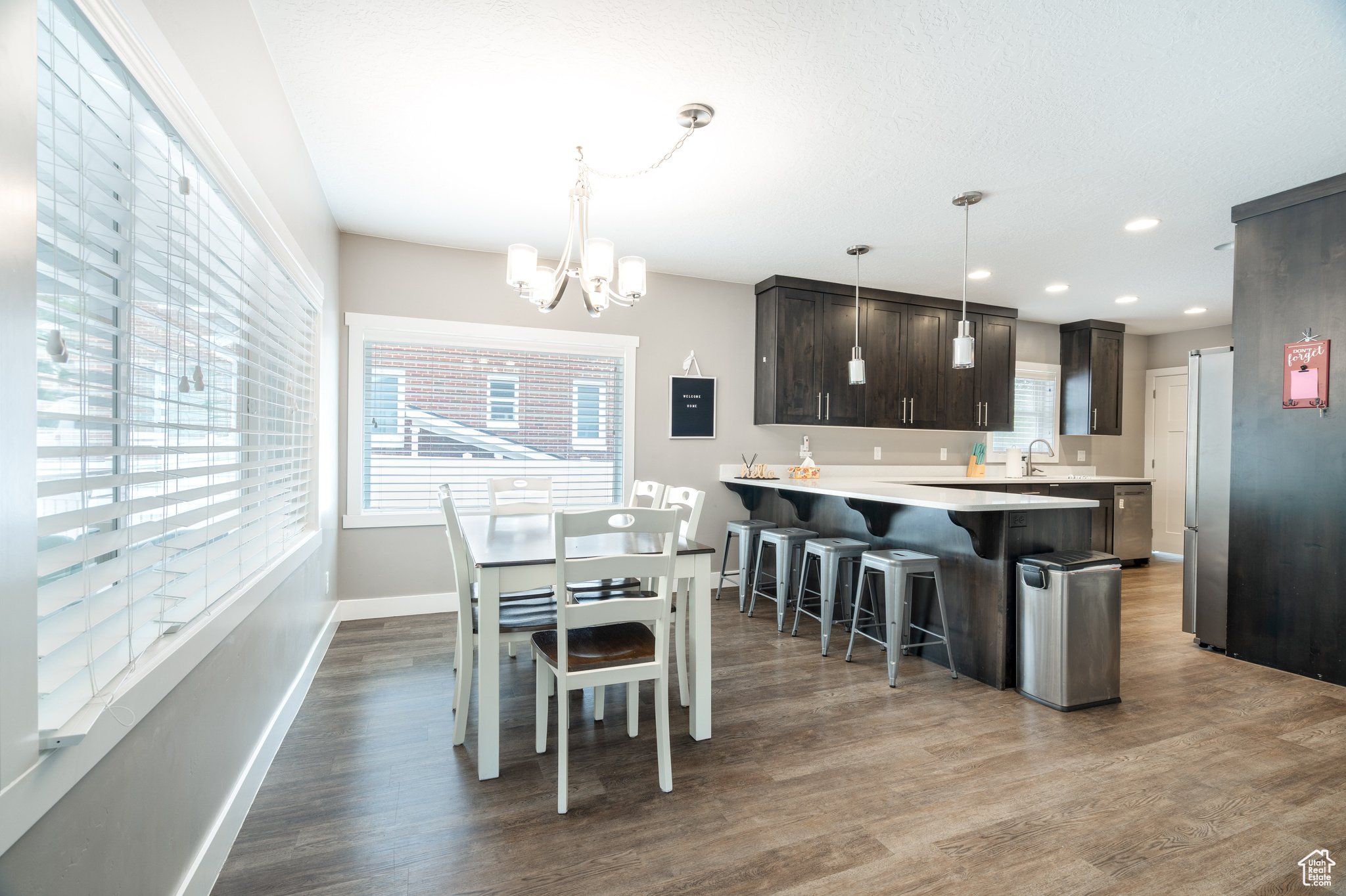 Modern kitchen and dining area with large windows, white and dark gray cabinets, a white dining table with chairs, a chandelier, pendant lights over the counter, and wood flooring.