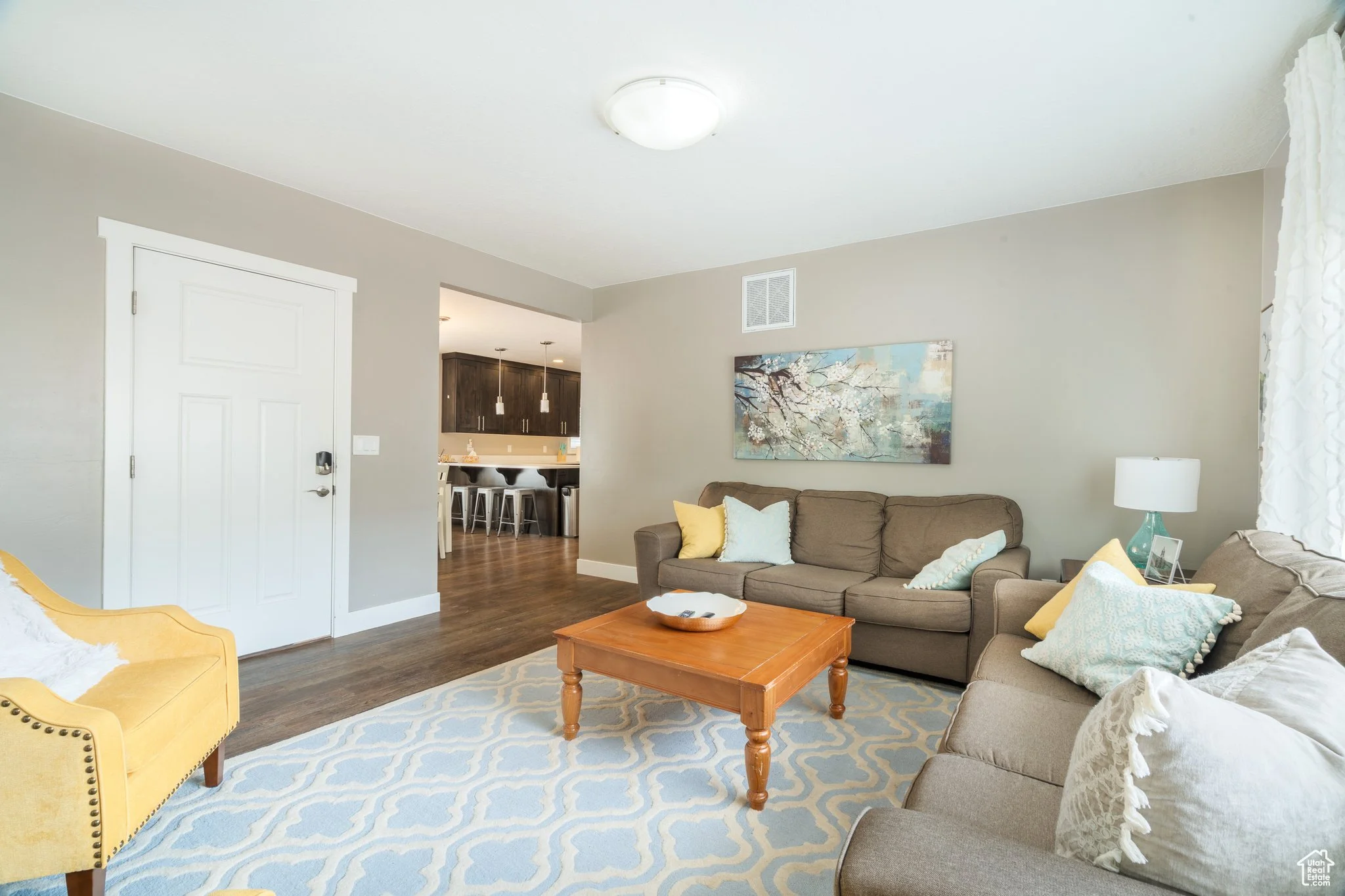 Living room with beige walls, a brown sofa with yellow, blue, and white pillows, a yellow armchair, a wooden coffee table, a blue and white patterned rug, a side table with a lamp, and a kitchen with dark cabinets and bar stools visible in the backgr