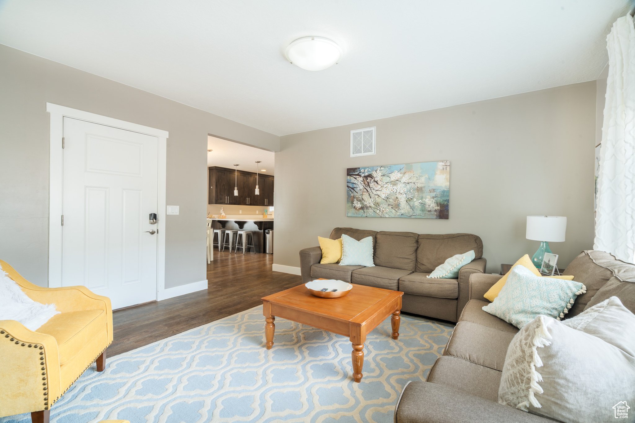 Living room with beige and yellow sofas, a wooden coffee table, and decorative pillows. A painting of blooming branches hangs on the wall, and a table lamp is on a side table. The room opens to a kitchen area with dark cabinets and a breakfast bar with stools.