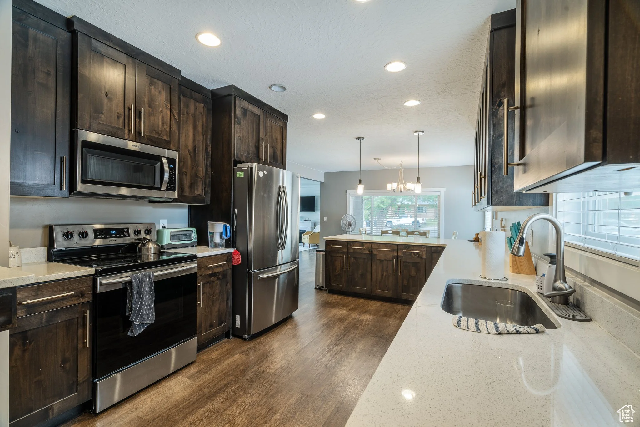 Modern kitchen with dark wooden cabinets, stainless steel appliances including a microwave, oven, and refrigerator, white countertops, and a window with blinds facing a patio area.