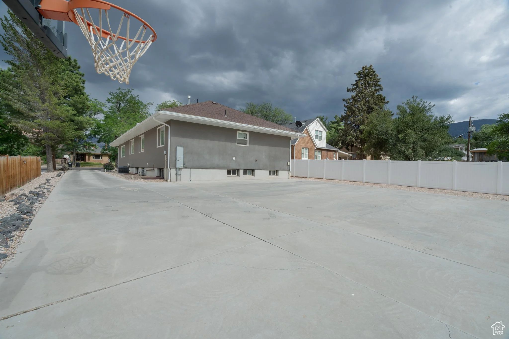 A backyard with a concrete patio, a white privacy fence, a basketball hoop, and a house under a cloudy sky.