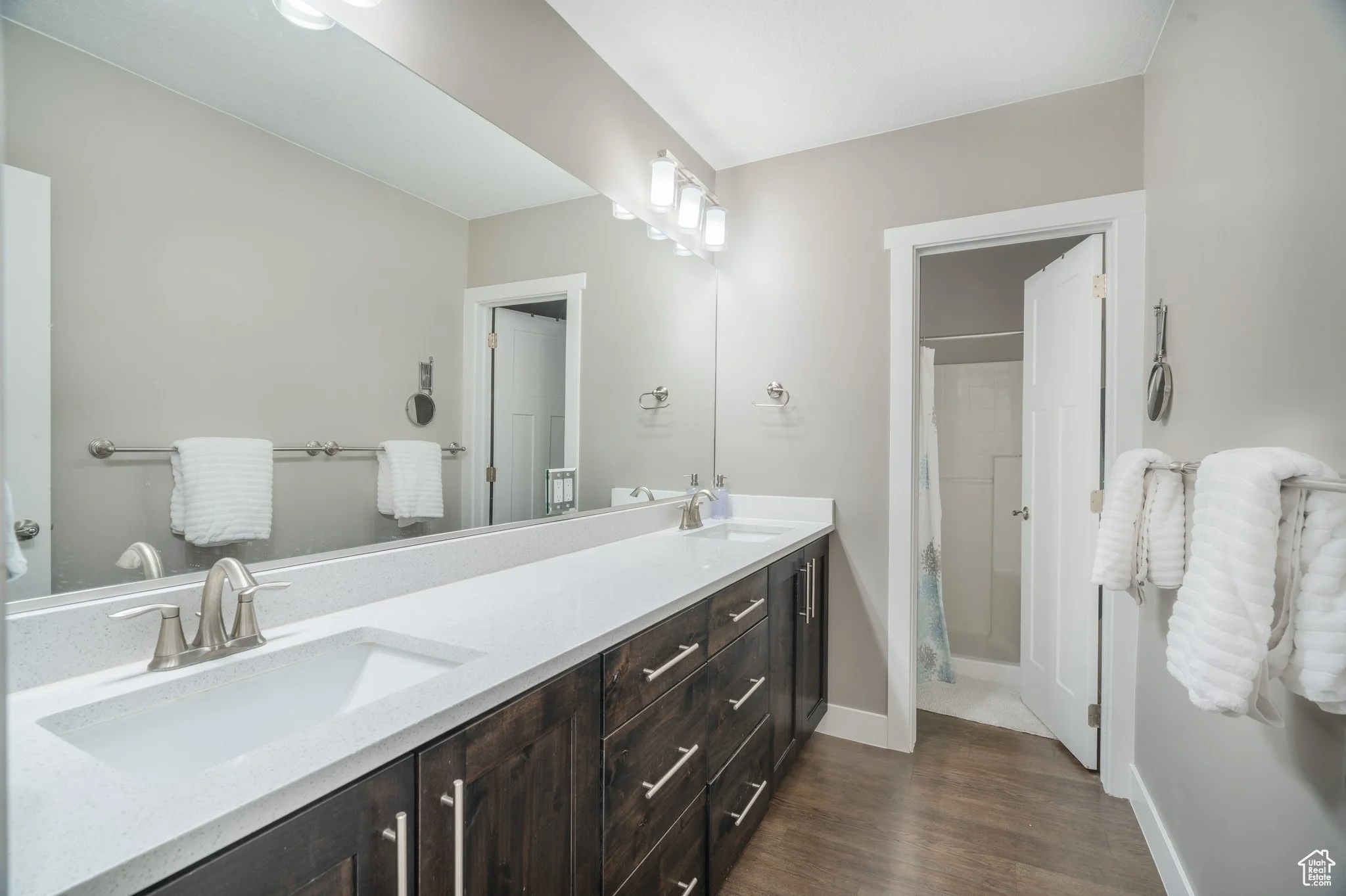 A modern bathroom with a double sink vanity, dark wood cabinets, and a large mirror. White towels hang on bars, and there is a small doorway leading to a shower or bathtub area in the background.