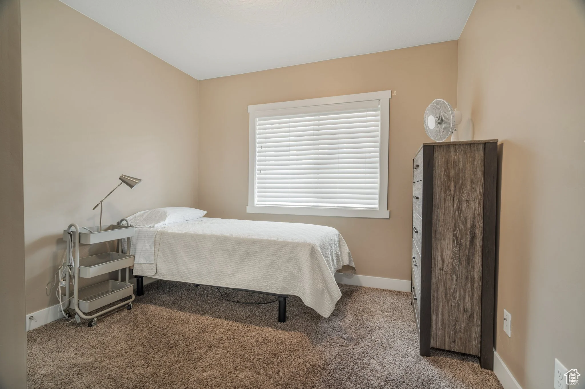 A small bedroom with beige walls, a single bed with white bedding, a window with closed white blinds, a tall dark wood dresser with a small fan on top, and a white rolling cart with a adjustable lamp and some cables.
