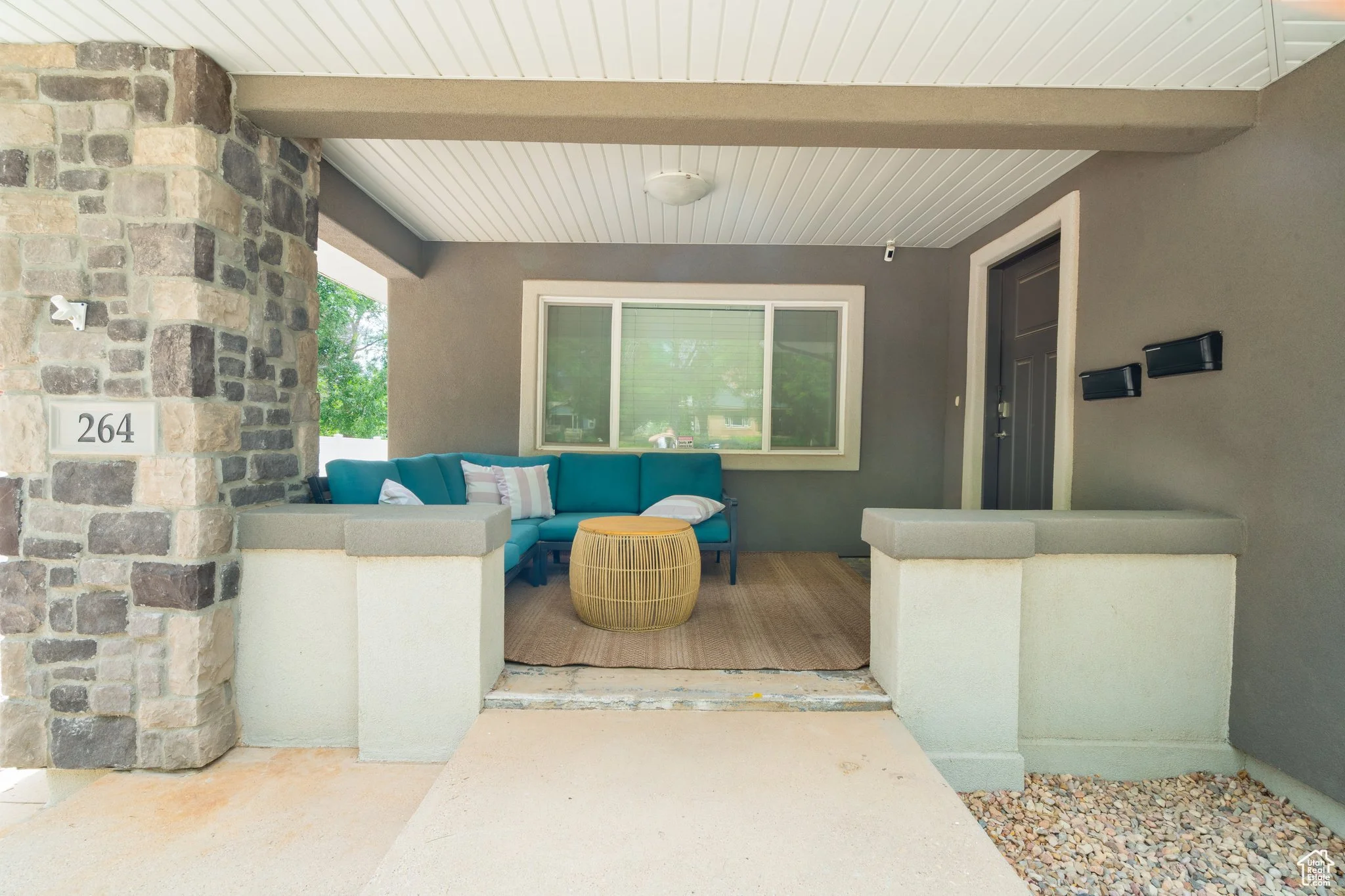 Front porch of a house with a cushioned blue outdoor sofa, a round wicker table, a window, and a black front door. The house has a stone and stucco exterior with house number 264.