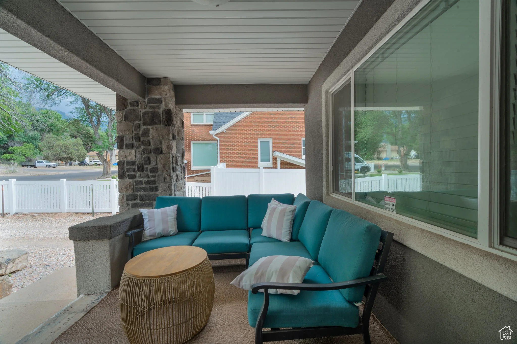 A covered porch with a blue sectional sofa and pillows, a round beige woven table, stone pillar, window, white fence, trees, and neighboring houses in the background.
